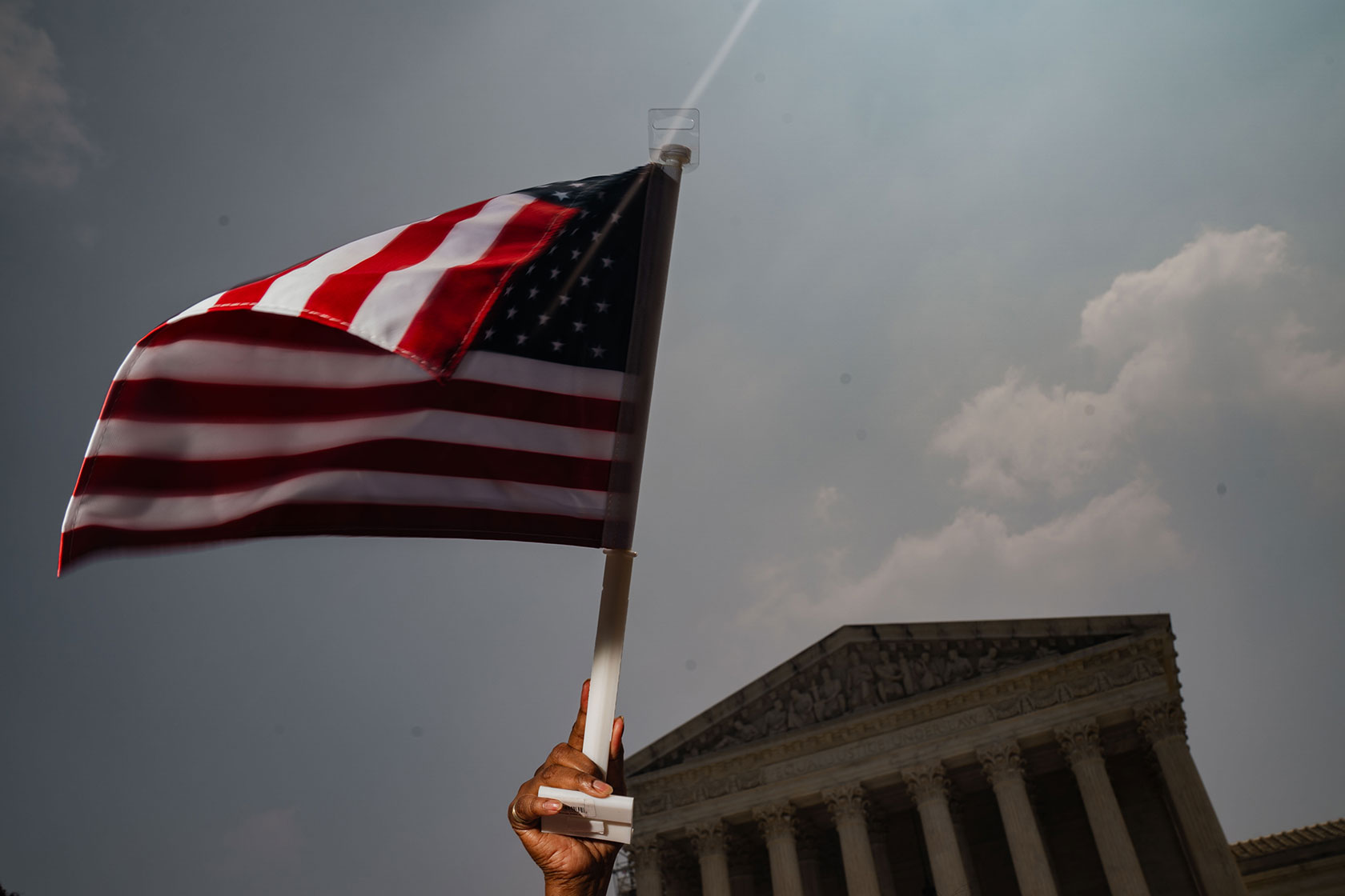 Photo shows a hand holding a small American flag, with the Supreme Court building against a cloudy sky in the background