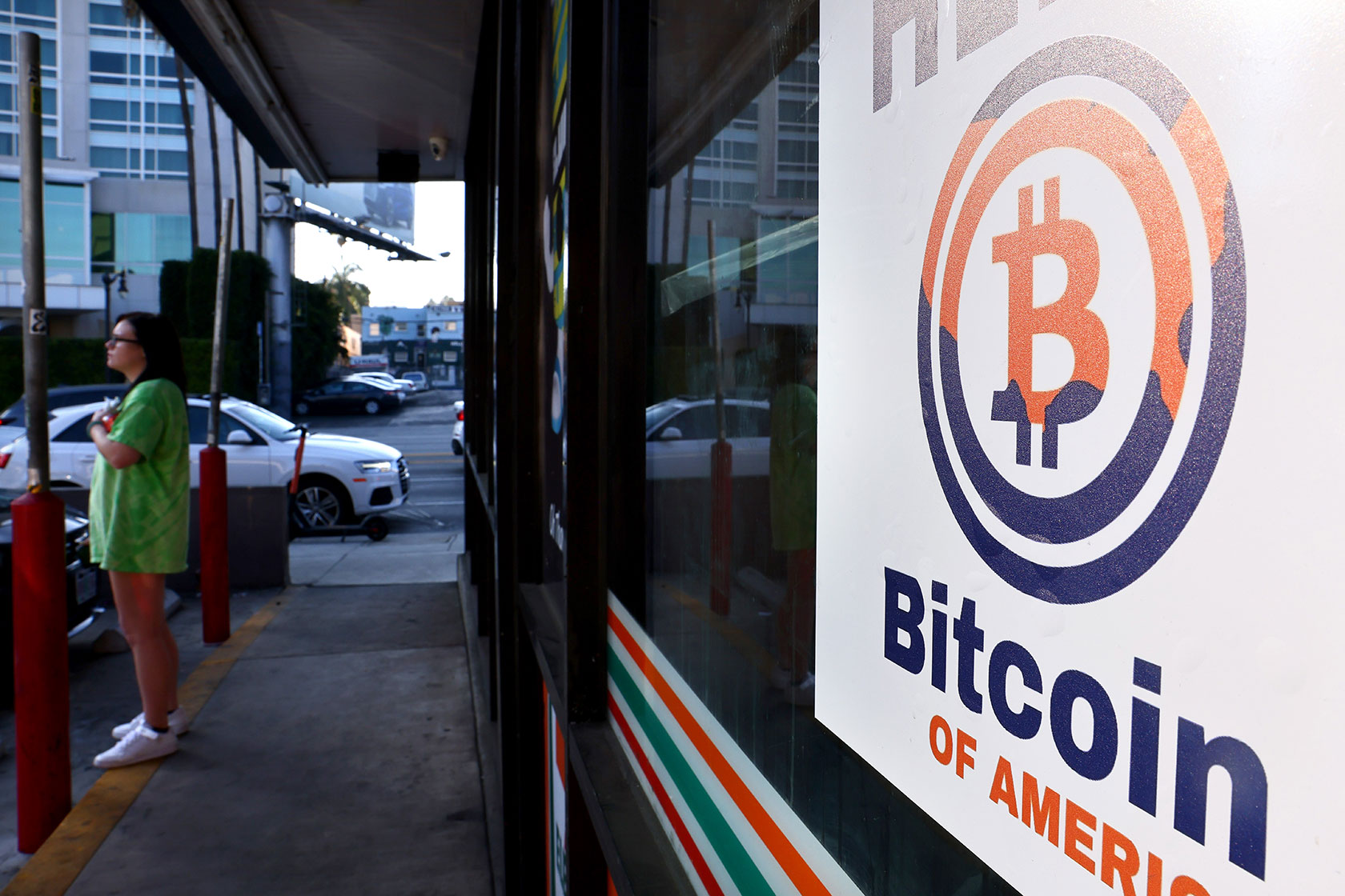 Photo shows a sign with the Bitcoin symbol in a storefront window, with a woman standing on the sidewalk in the background.