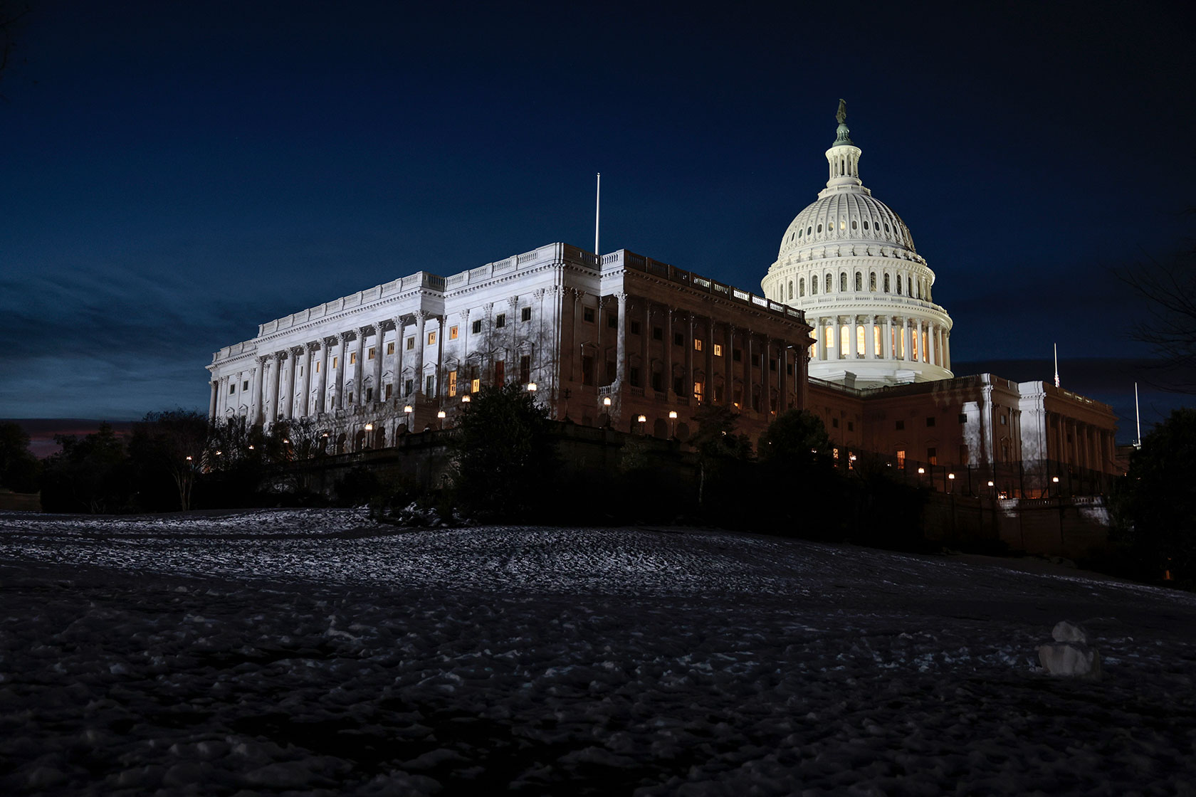 Photo shows the Capitol building glowing white against a dark blue sky, with snowy grounds in the foreground