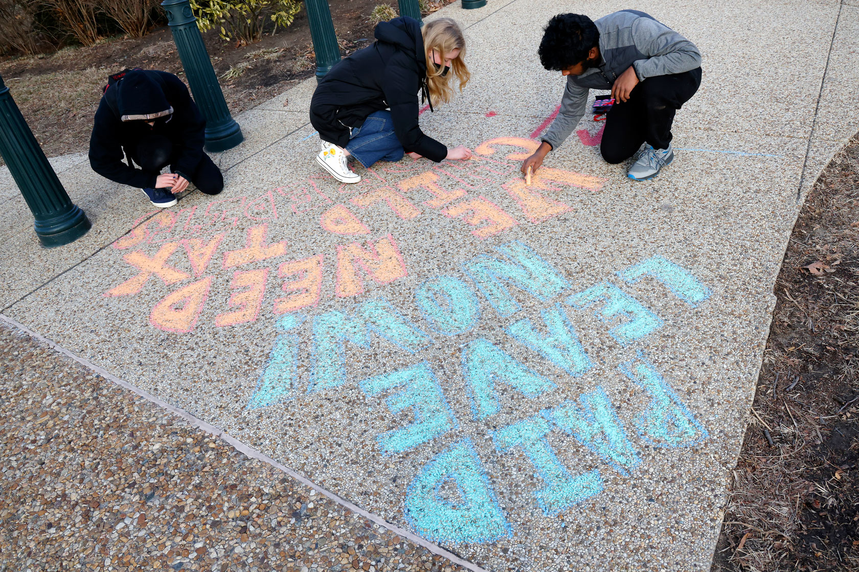 Three people crouch on the sidewalk to write a message in blue and orange chalk.