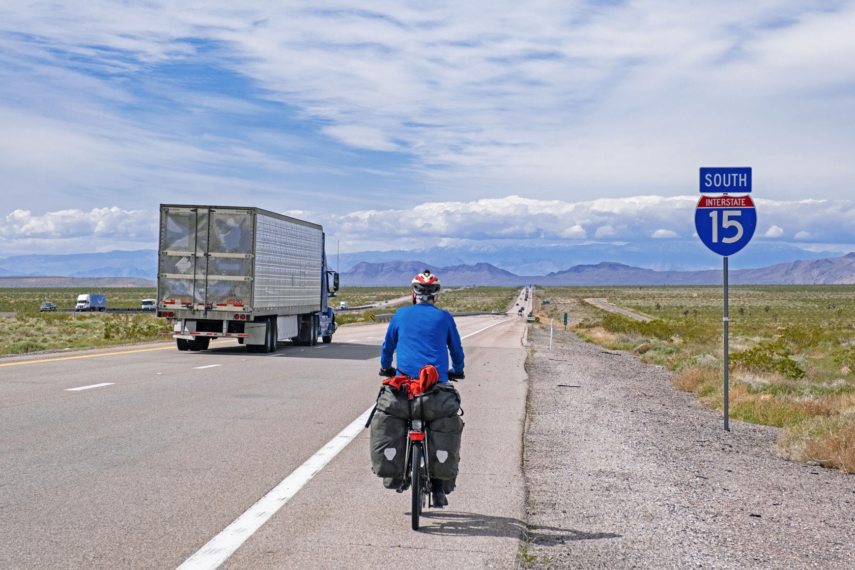 A cyclist is seen alongside a truck on a long strand of road with mountains and clouds in the background.