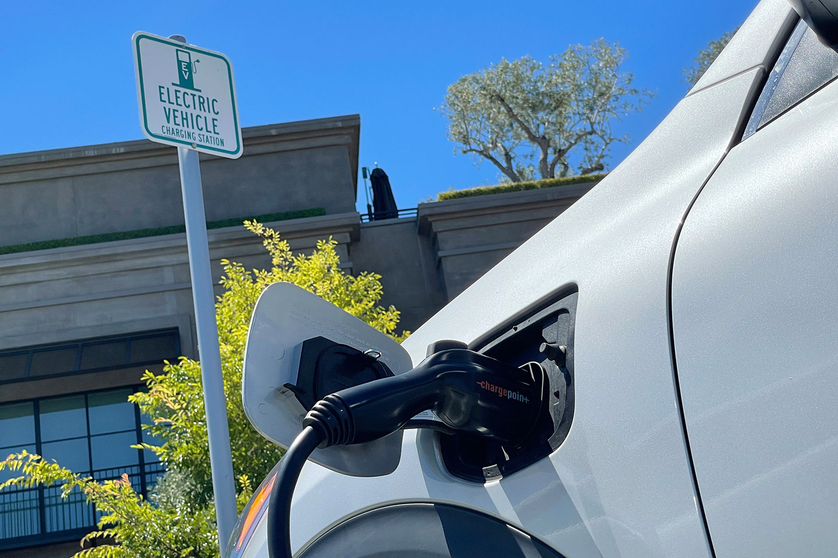 Photo shows a close-up of a white car hooked up to a charging station, with a sign reading 