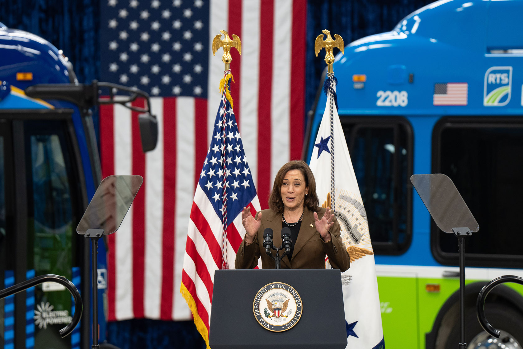 Kamala Harris is seen at a podium in front of two flags and two electric buses.