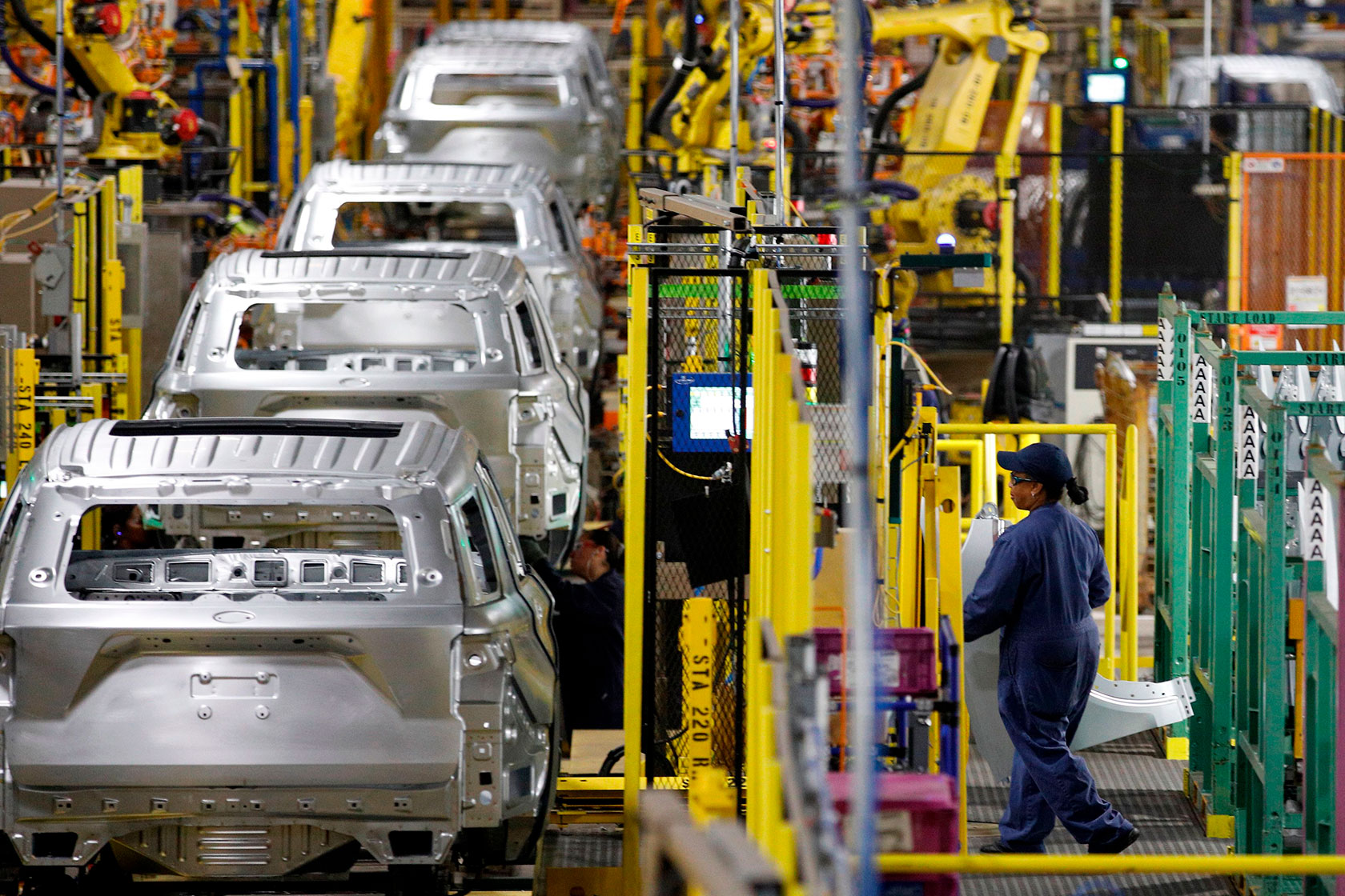 Photo shows a row of partially assembled cars with a worker standing on the side