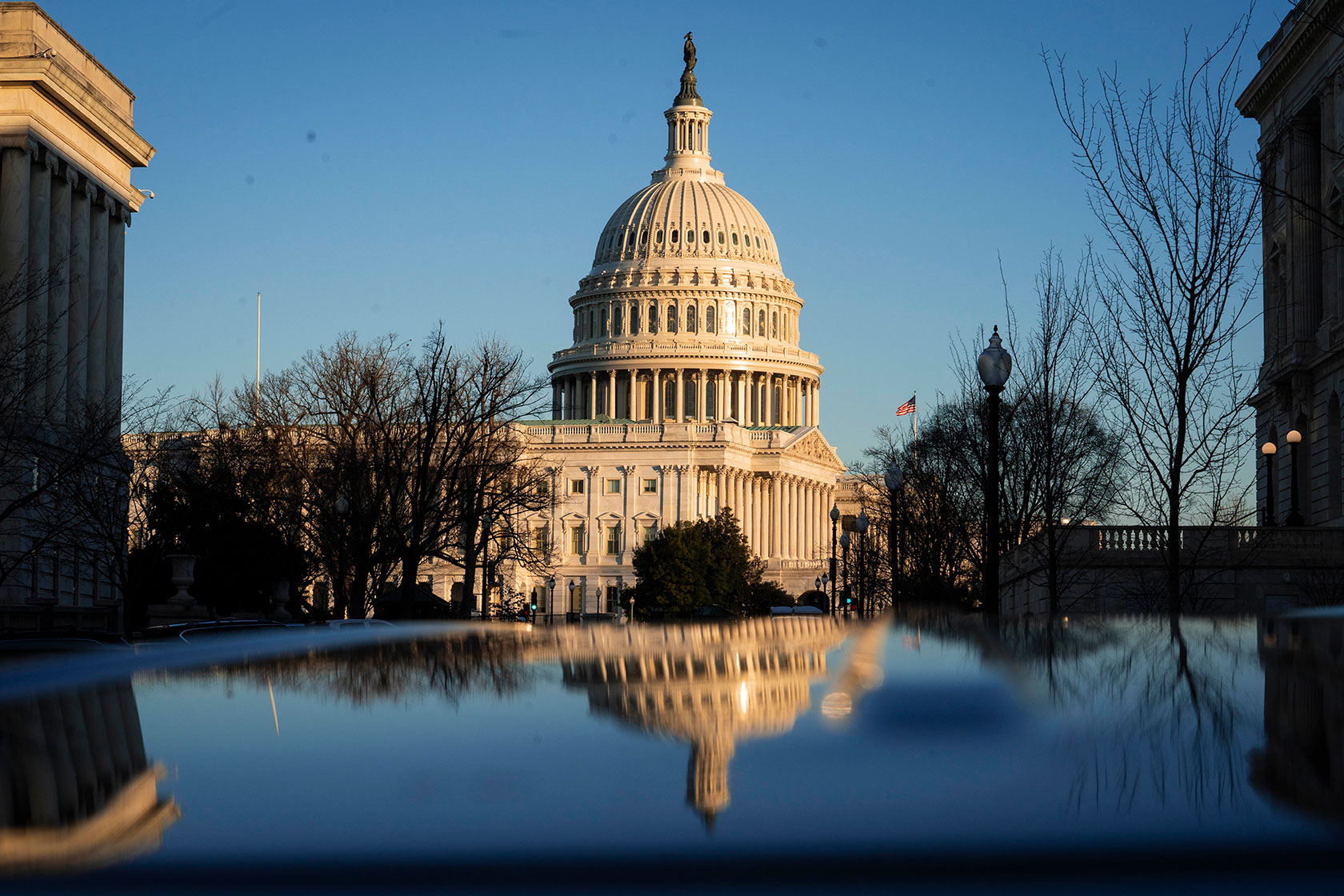 Photo shows a view of the Capitol building against a blue sky, partly reflected in a shiny surface in the foreground