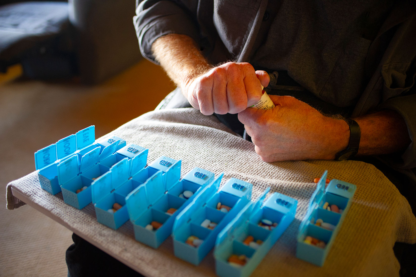 Photo shows a closeup of a person's hands opening a bottle of pills, with a blue pill organizer sitting in front of them