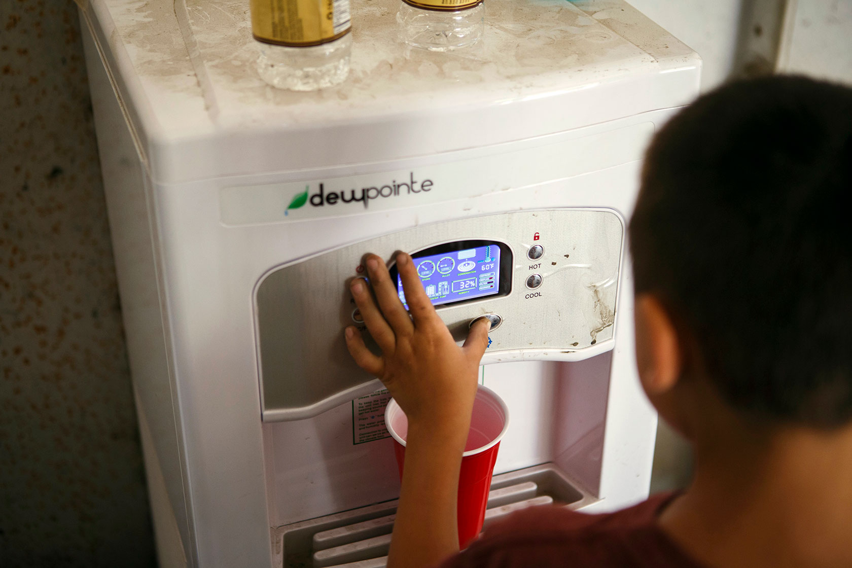 Photo shows the back of a young boy wearing a red shirt filling up a red plastic cup with water from a dispenser