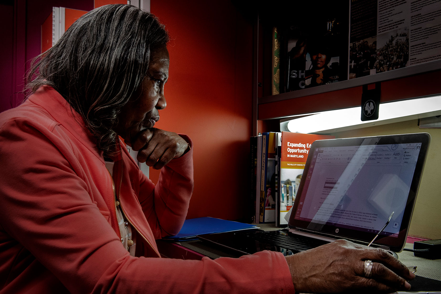 Phot shows a woman in a pink jacket sitting in front of her computer looking thoughtfully at the screen