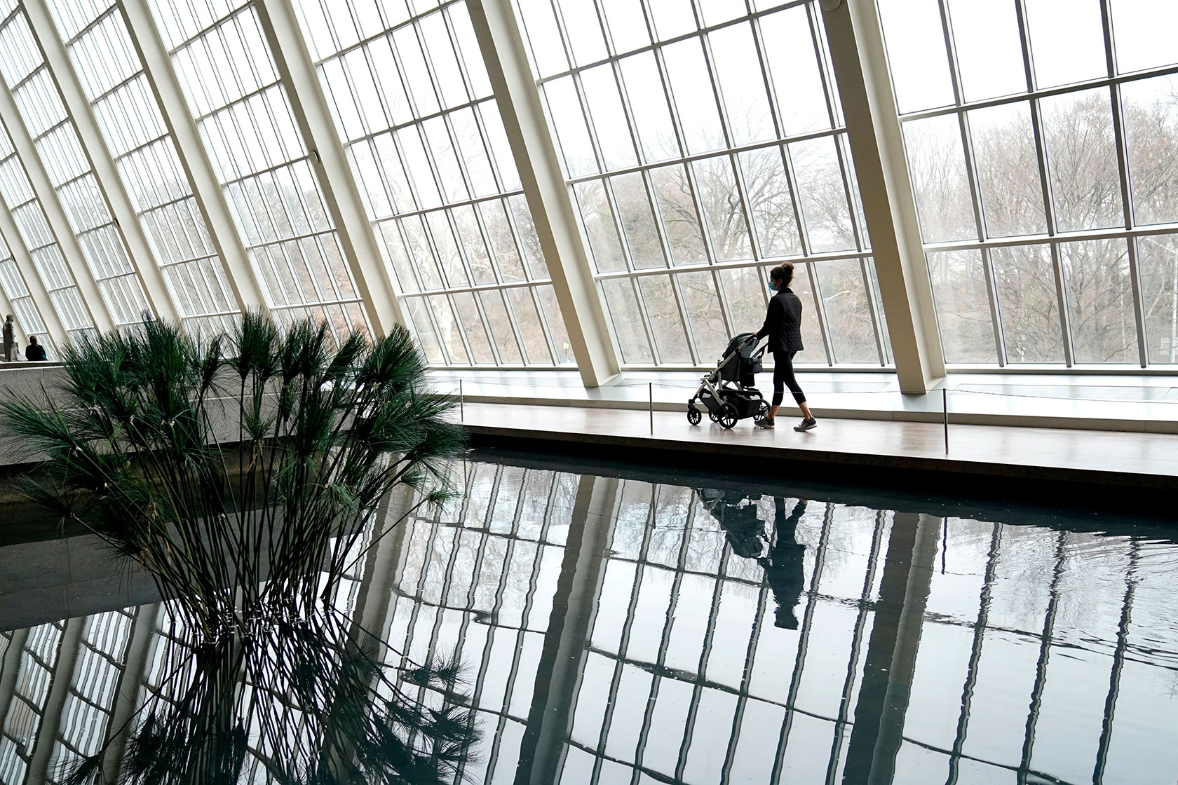 A woman pushing a stroller walks through an atrium with light pouring in and a small pool reflecting plant life