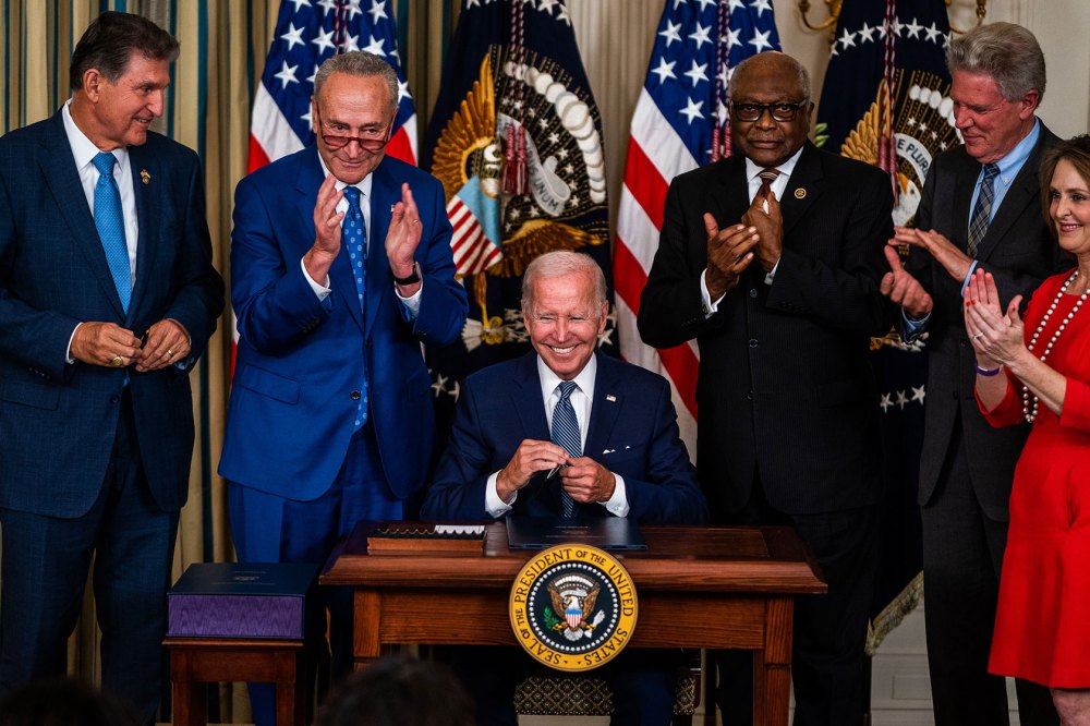 Photo shows Joe Biden sitting at a desk, smiling, surrounded by members of Congress standing around him.