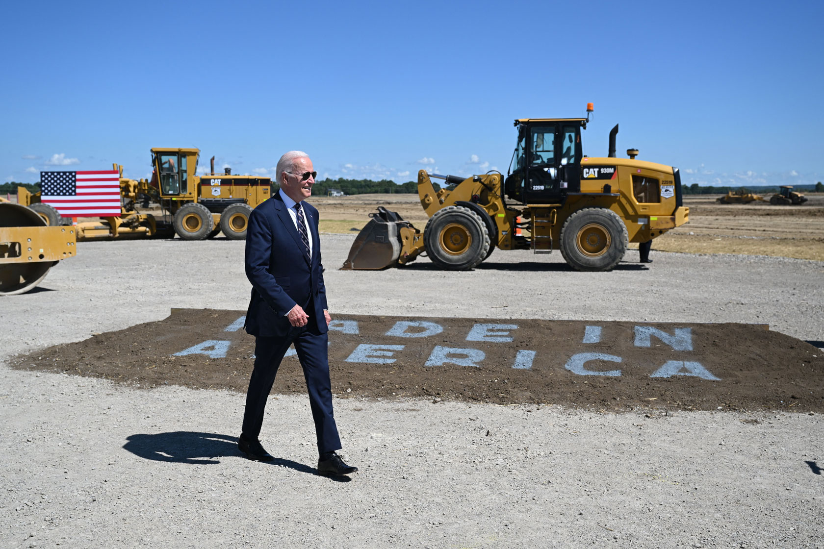 President Joe Biden is seen walking in front of tractors, with an American flag in the background.