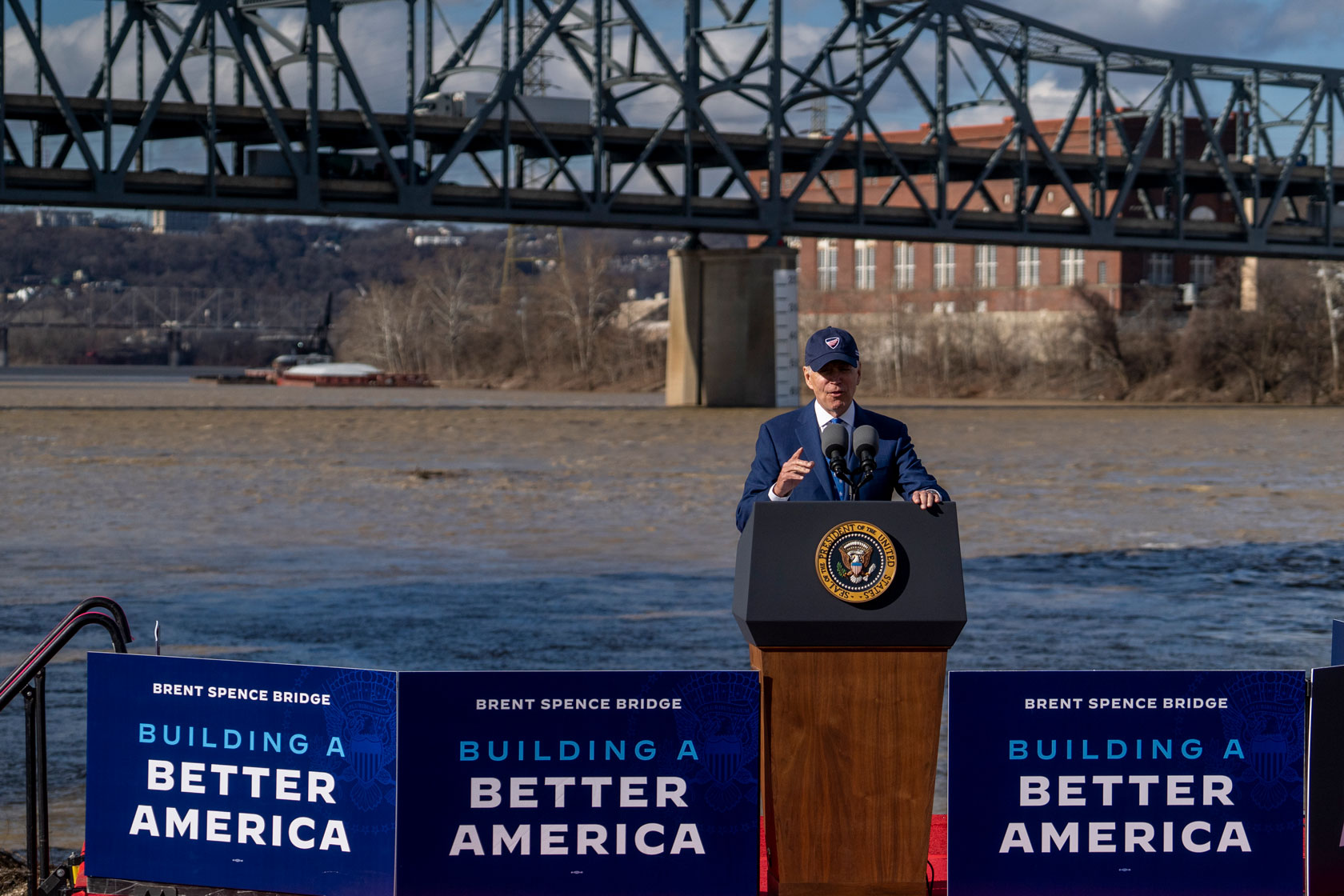 President Joe Biden is seen at a podium in front of a river with a bridge in the background.