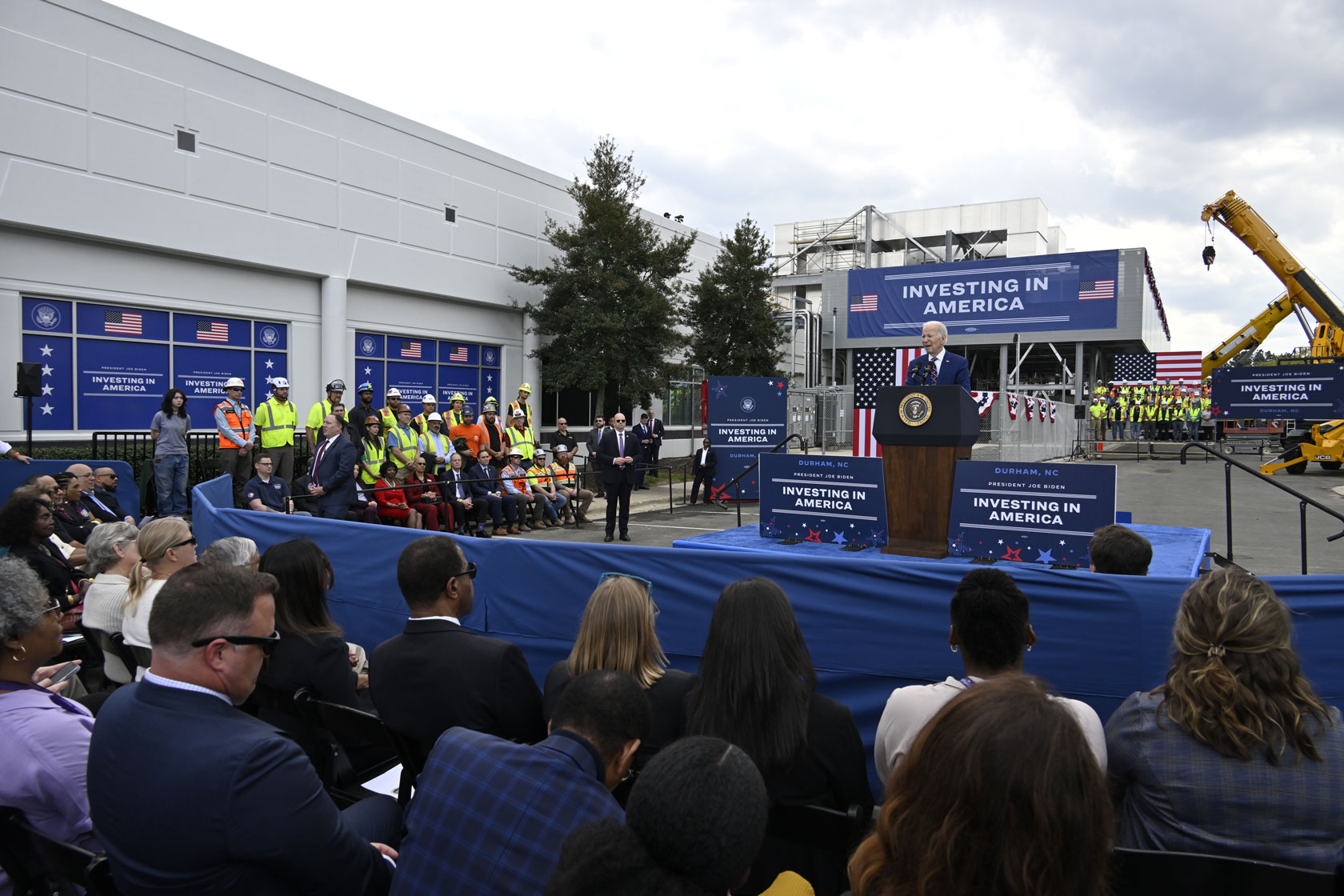 President Joe Biden speaks outside of a building at a podium in front of a crowd including construction workers.