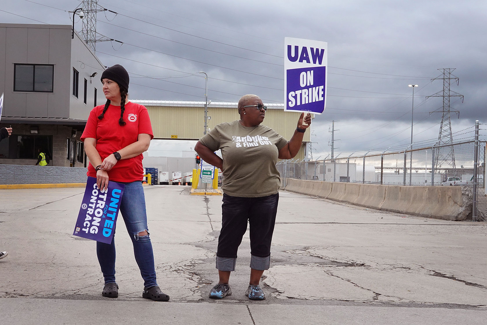 Photo shows two women holding signs in front of a grey building. One sign says 