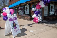 An "open" sign with balloons attached to it is seen in front of a brick storefront decorated with a balloon arch.