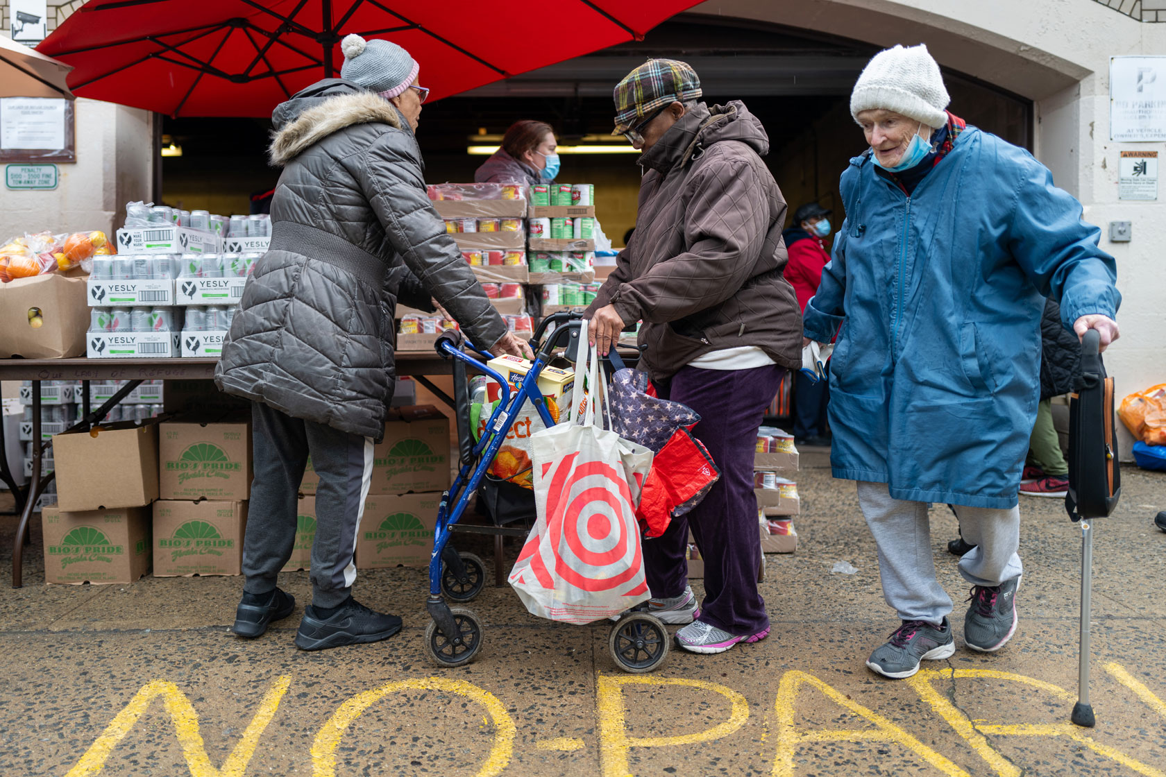 Three women are seen under a red umbrella in front of a table that has stacks of cans on it.