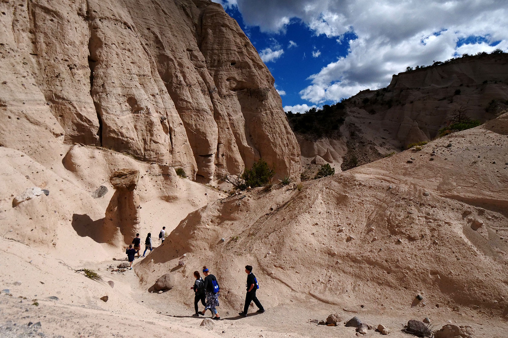 Photo shows a small group of people hiking among tall, pastel orange rock formations against a blue sky with some puffy white clouds