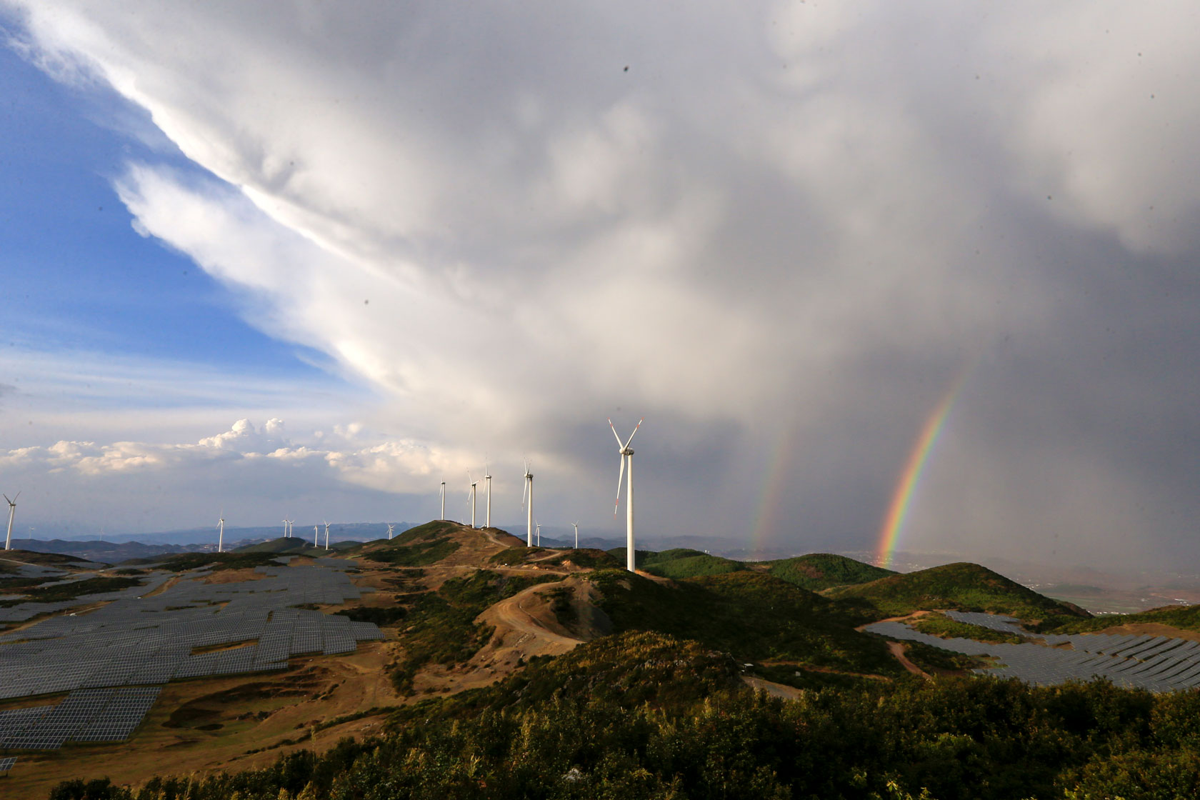 A line of windmills is seen at the top of a hill with solar panels in a valley to the left and a rainbow to the right.
