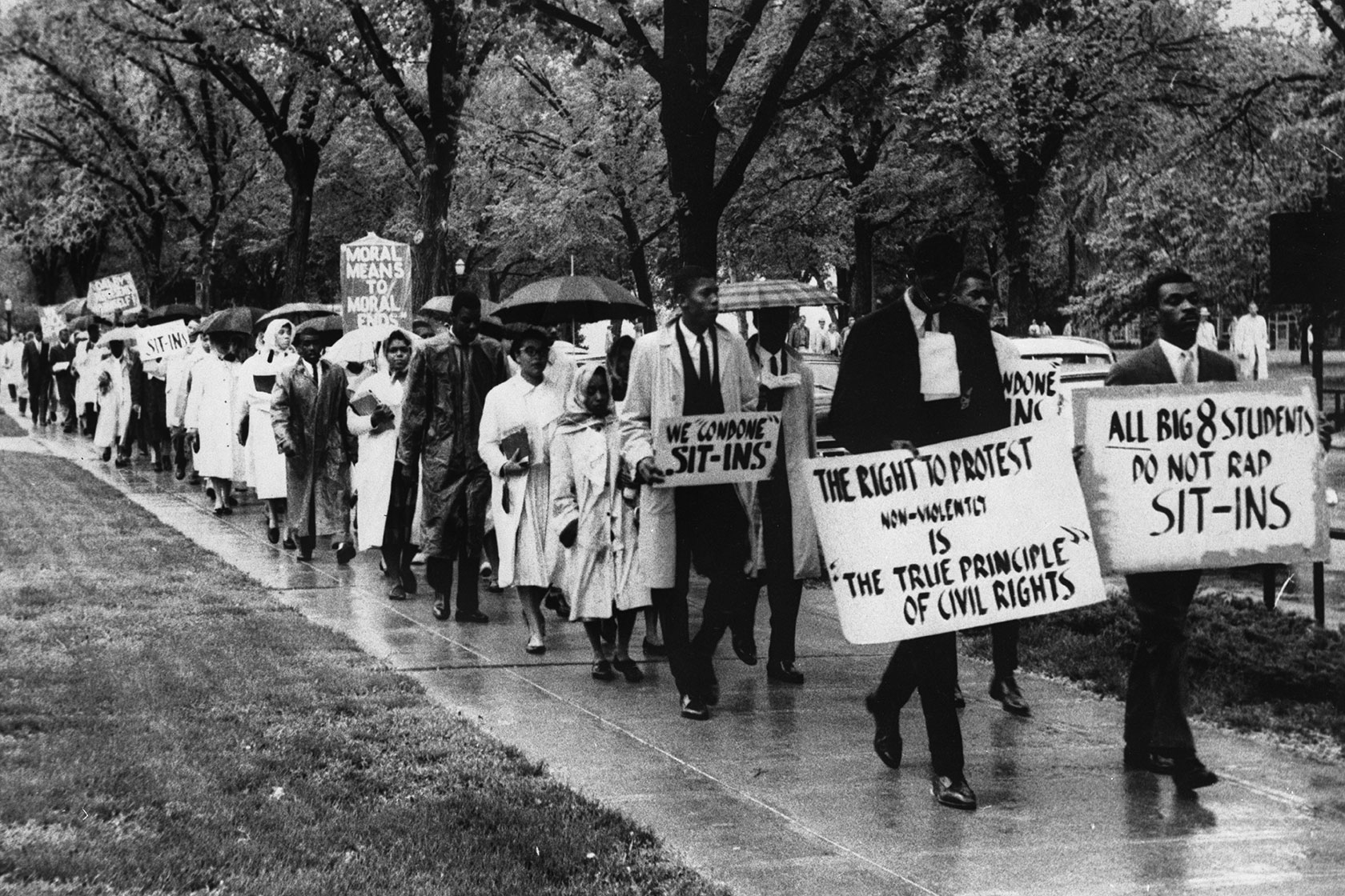 Photo shows a historical black and white image portraying a group of Black students marching through a treed section of a university campus holding signs that read 