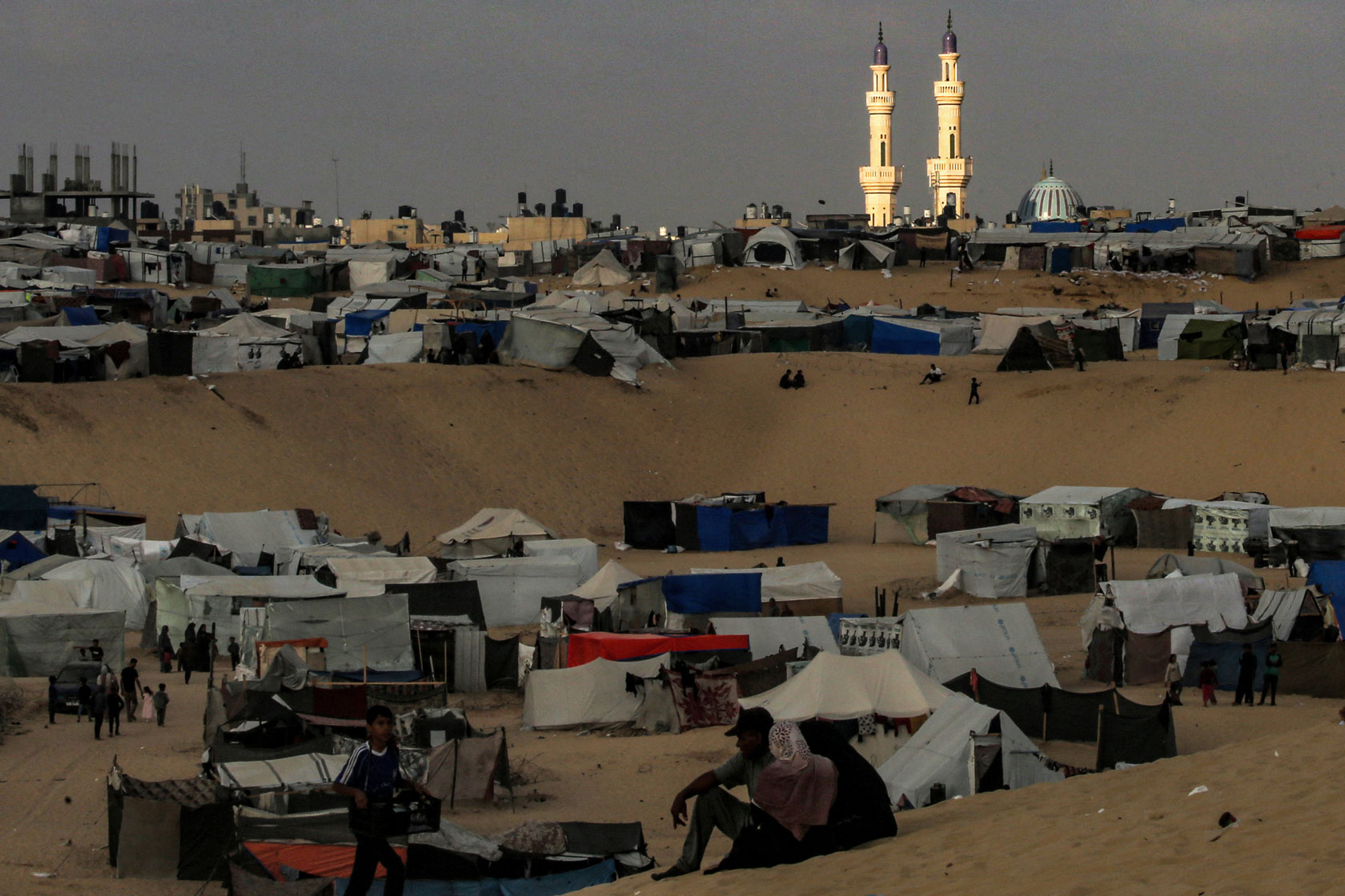A cluster of tents set up on top of rolling sand dunes is seen with the al-Taiba mosque at sunset in the background.