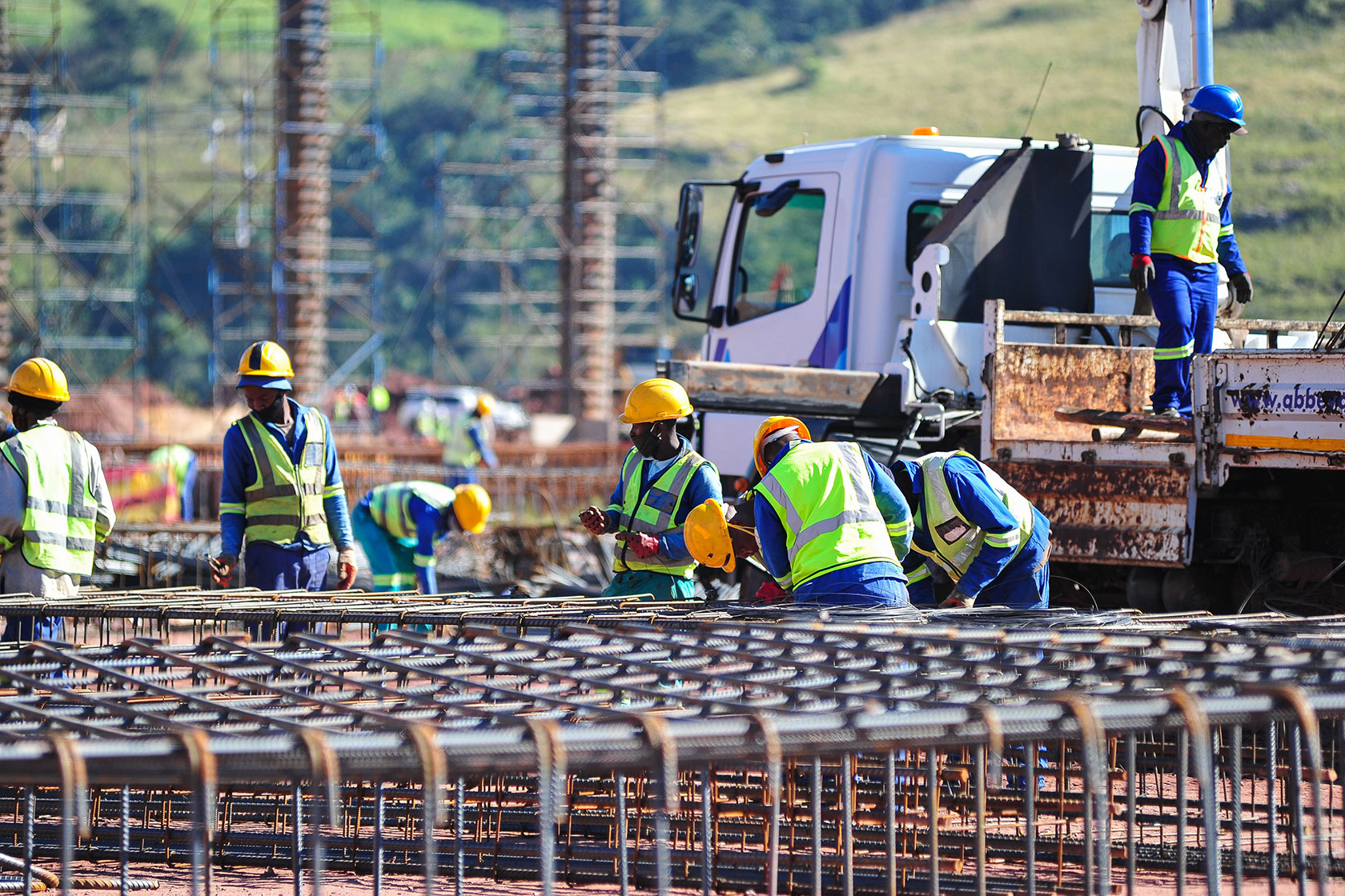 Photo shows workers in bright green construction vests and yellow hats setting up a metal frame with a truck in the background