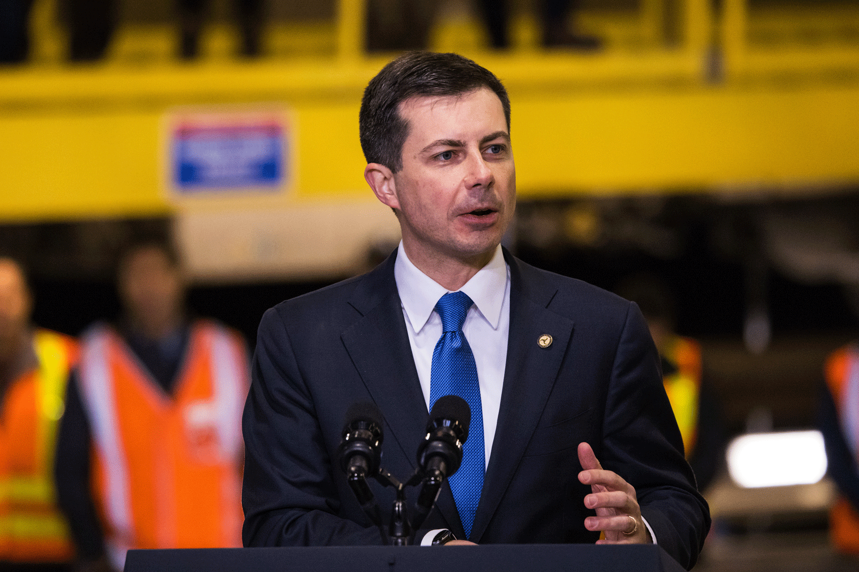 Pete Buttigieg is seen speaking behind a podium with construction workers in the background.