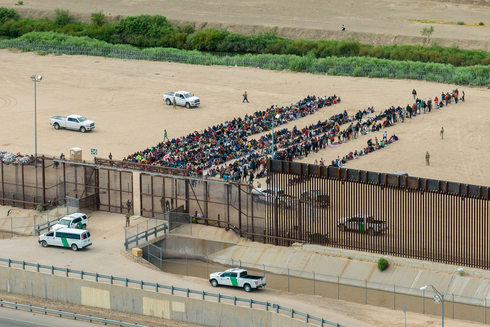 Photo shows groups of people sitting on sand behind a large fence, with white government trucks nearby