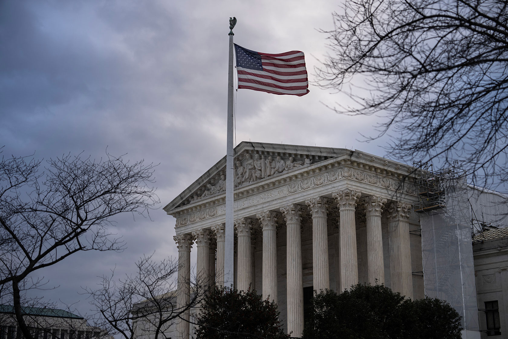 Photo shows a partial view of the front of the Supreme Court building against a cloudy sky, with the American flag flying in front