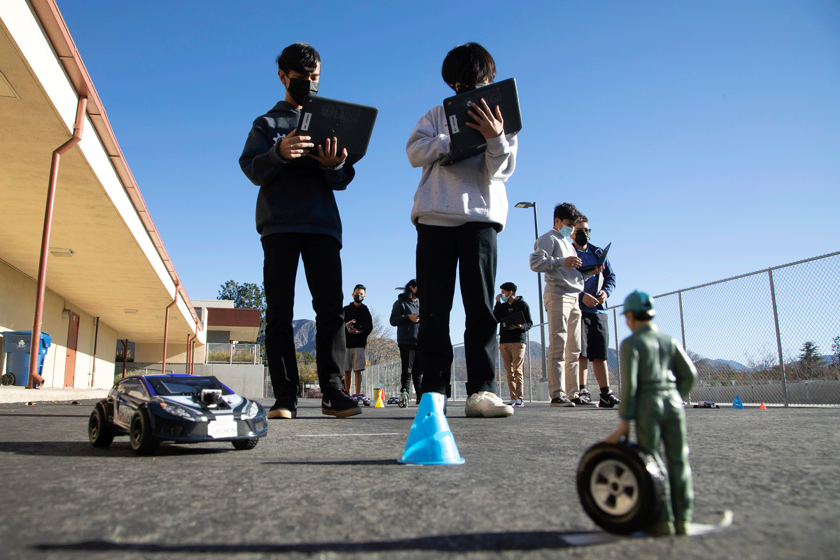 Two students stand outside with laptops in front of a toy person, cone, and robotic car.