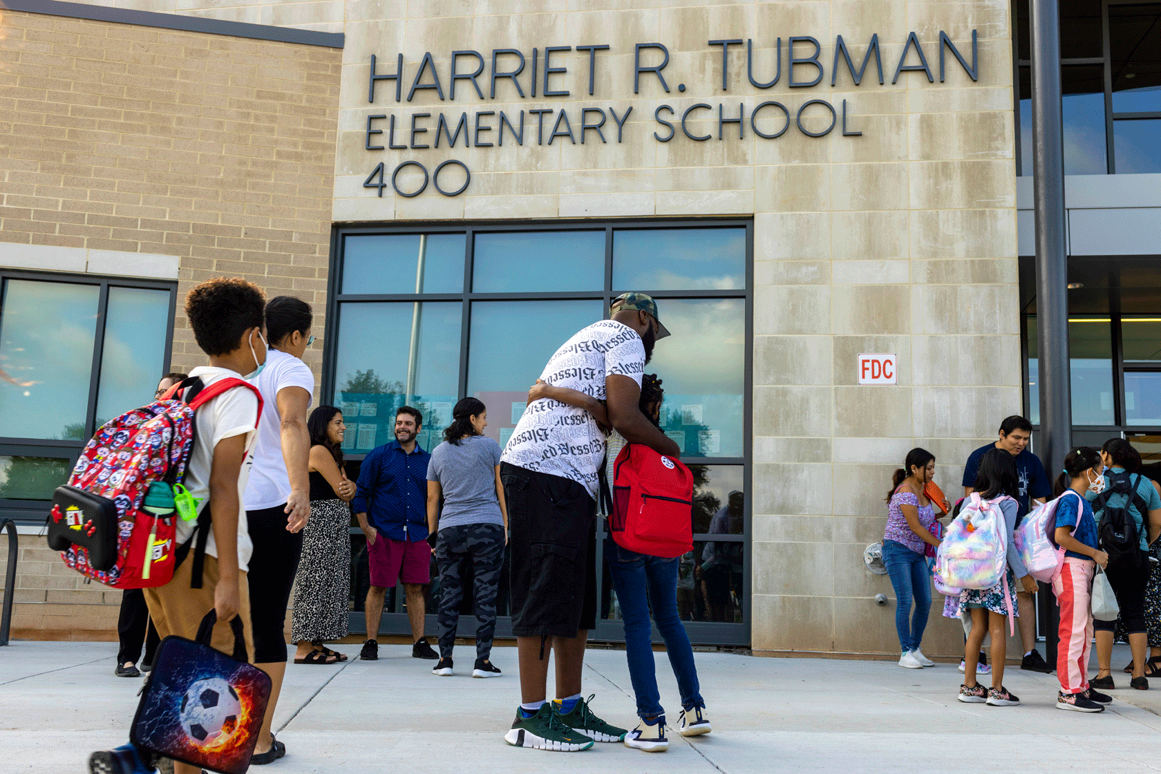 A parent and child are seen hugging outside of a school with other students waiting in the background.