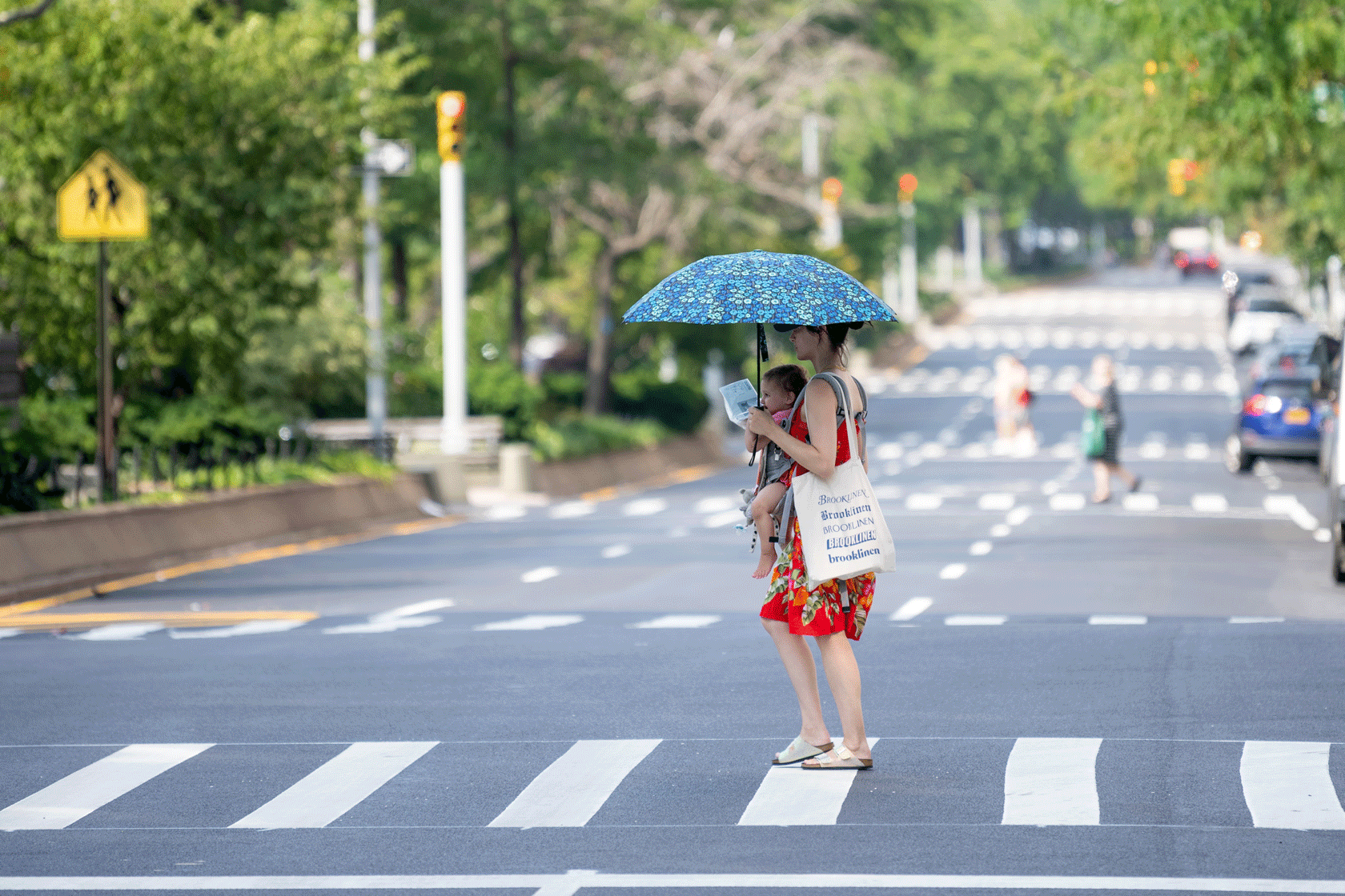 A mother carrying her baby in a baby carrier and holding an umbrella is seen crossing the street.