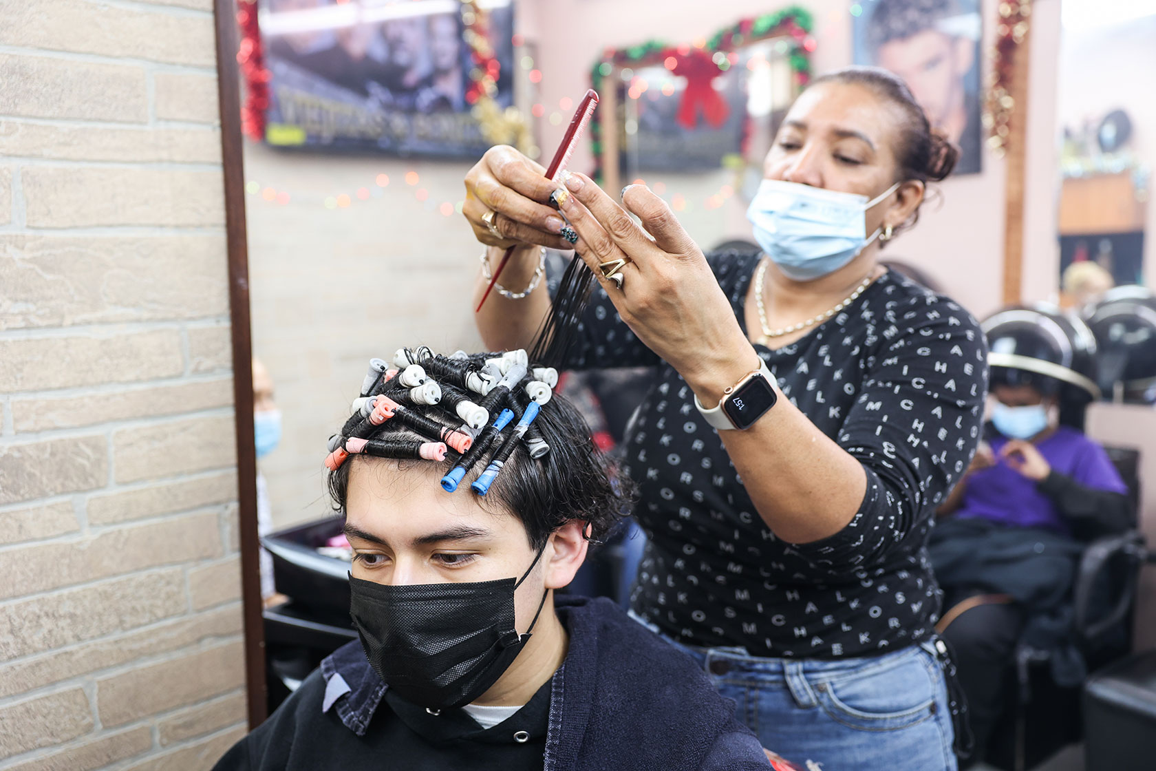 Photo shows a masked woman doing a teenage customer's hair reflected in a mirror