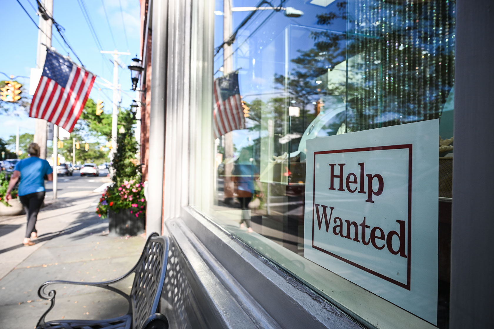 Help wanted sign in storefront window; person walking past near American flag