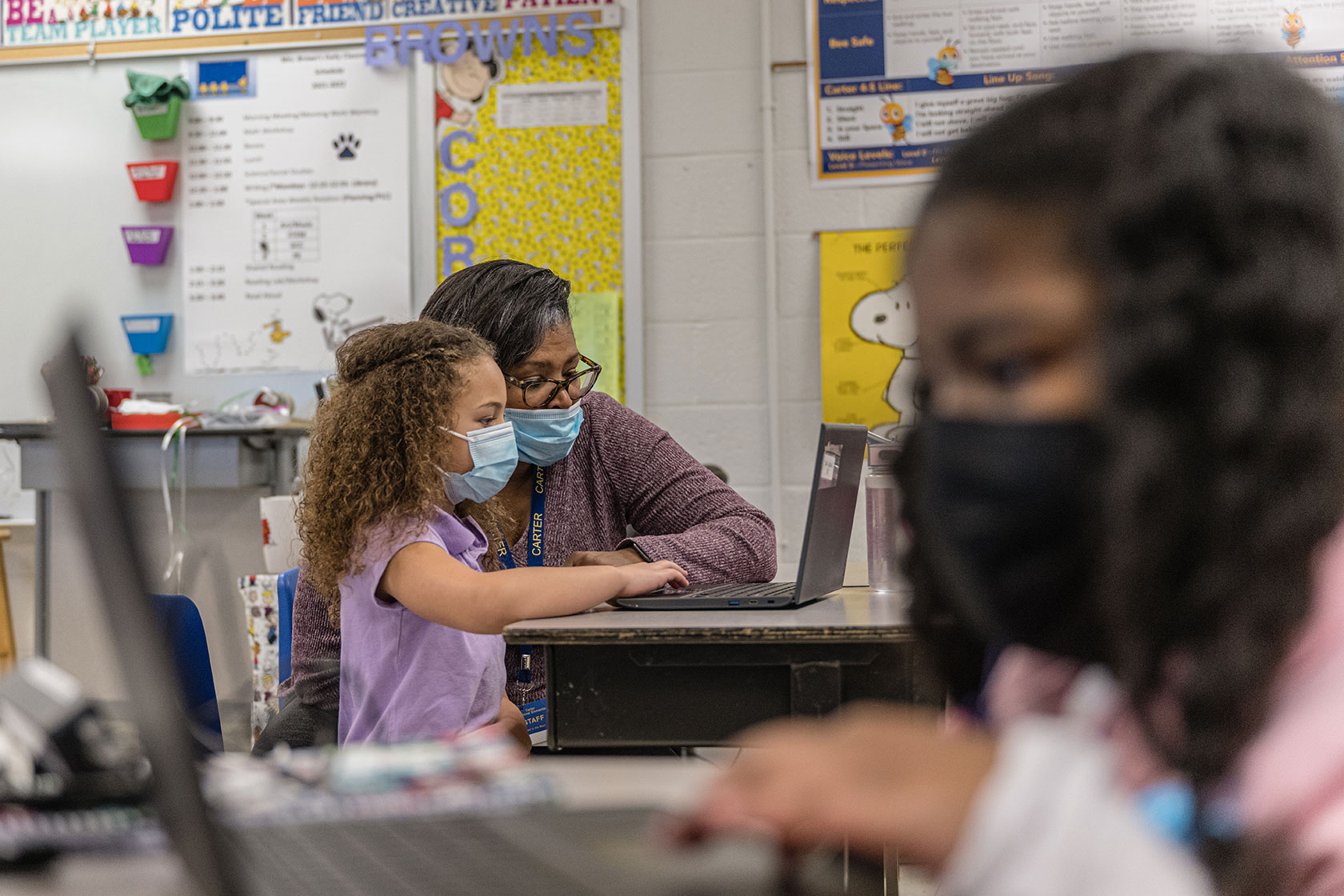 A teacher sits at a laptop computer with a student