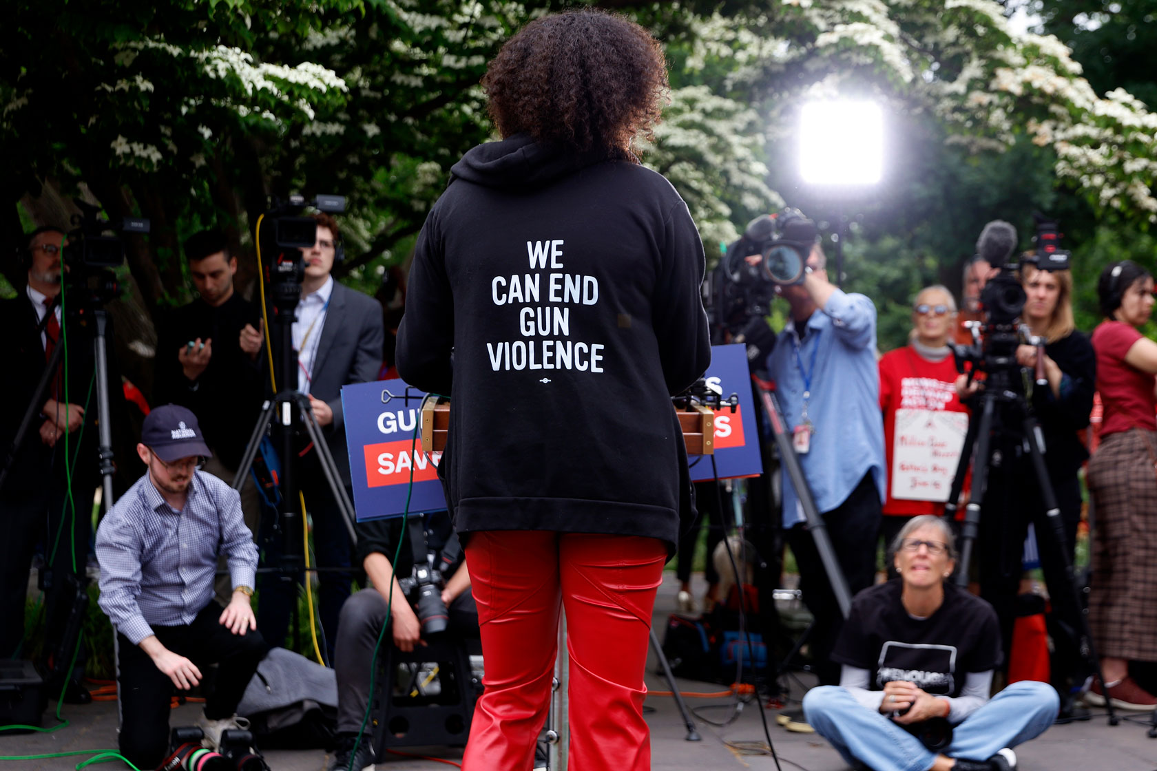 A woman in a black sweatshirt stands before media cameras. The words on her sweatshirt say, 