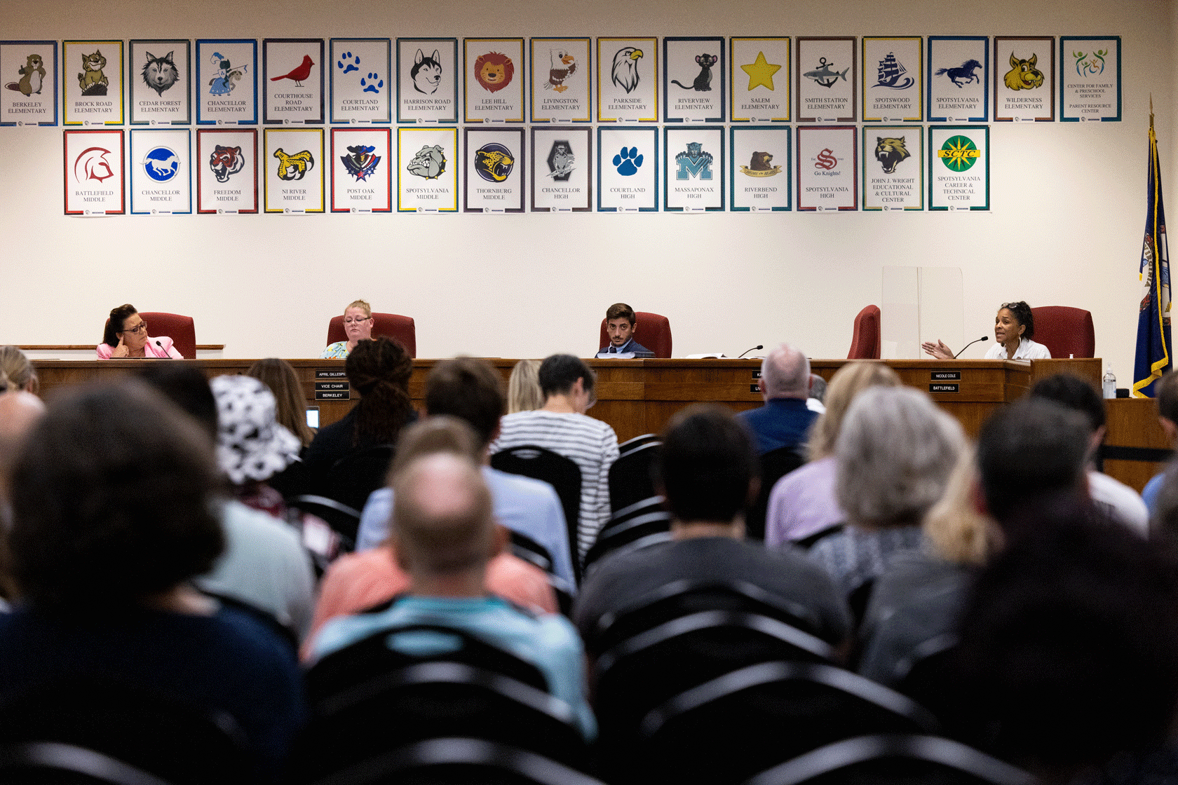 School board members are seen at a dais below framed pictures on the wall with audience members' heads in the foreground.