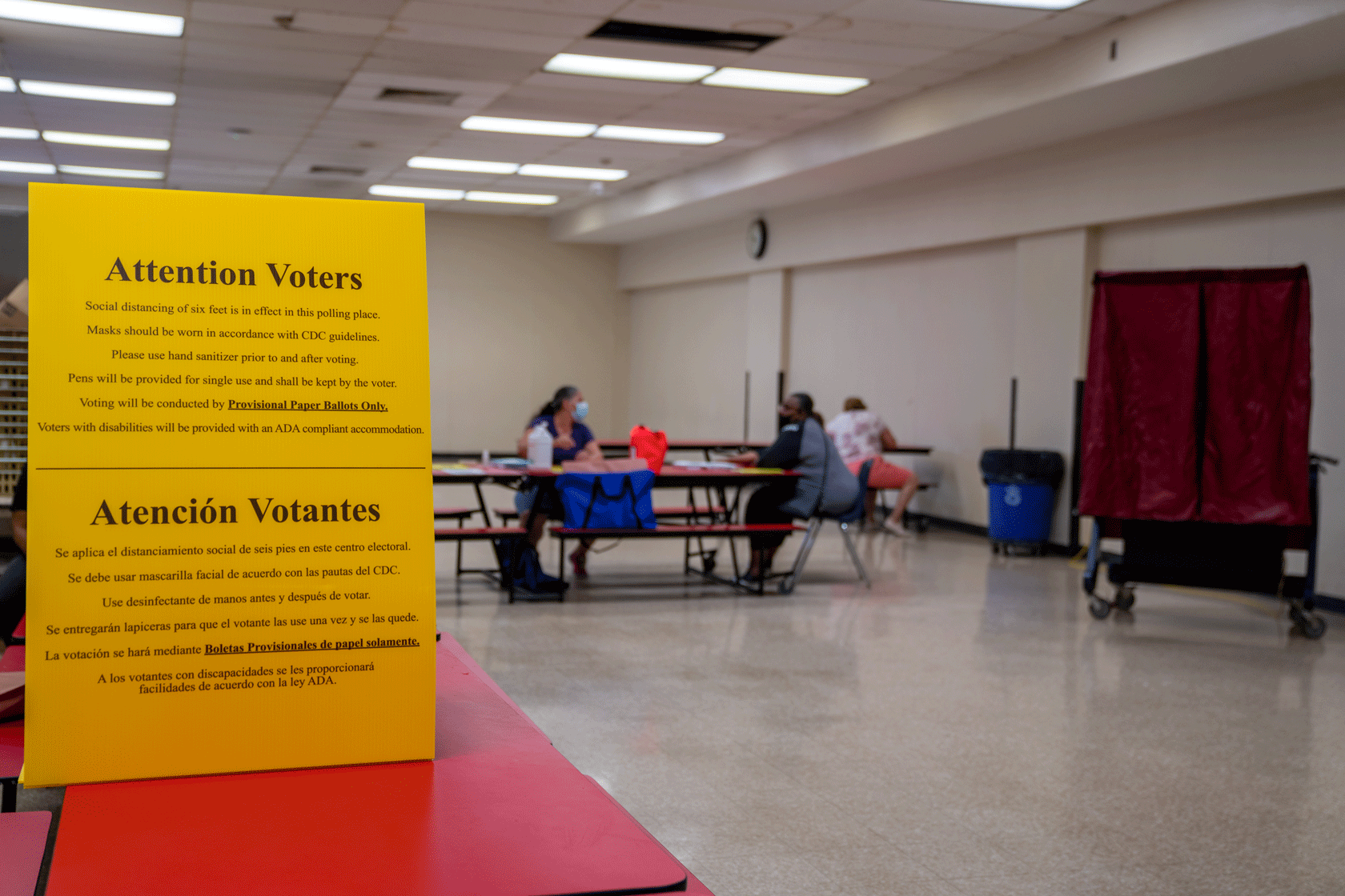 A sign is seen, with a red voting booth and some people sitting at red tables in the background.