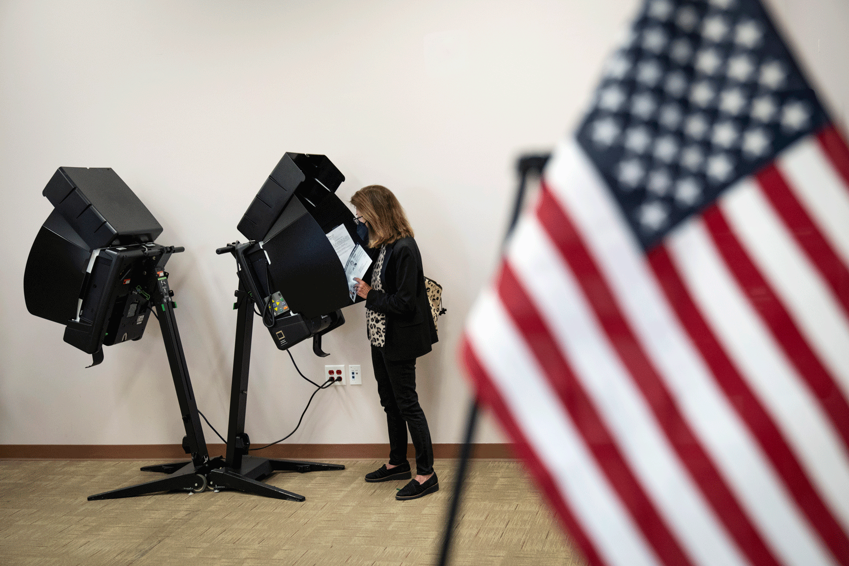An American flag is seen in the foreground while a voter stands at a polling booth in the background.