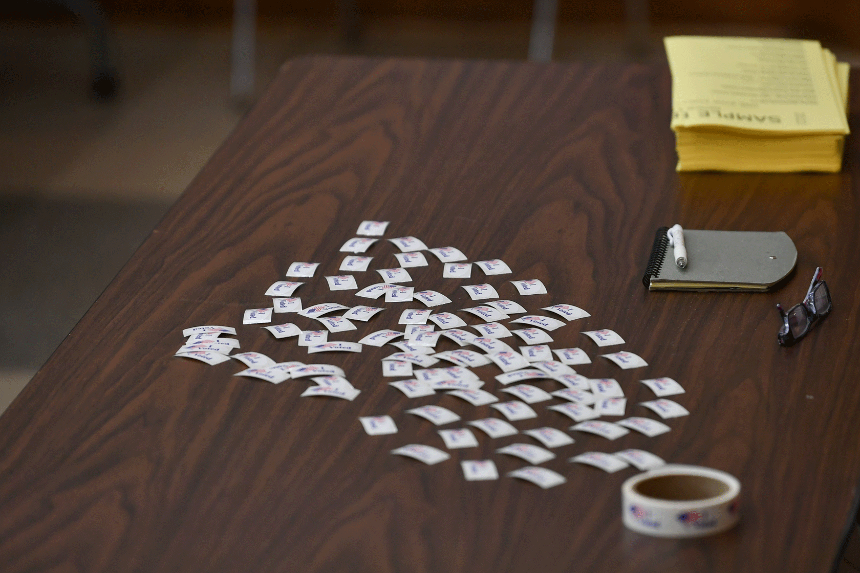 A table with scattered stickers, a notebook, a pair of glasses, and a stack of yellow flyers is seen.
