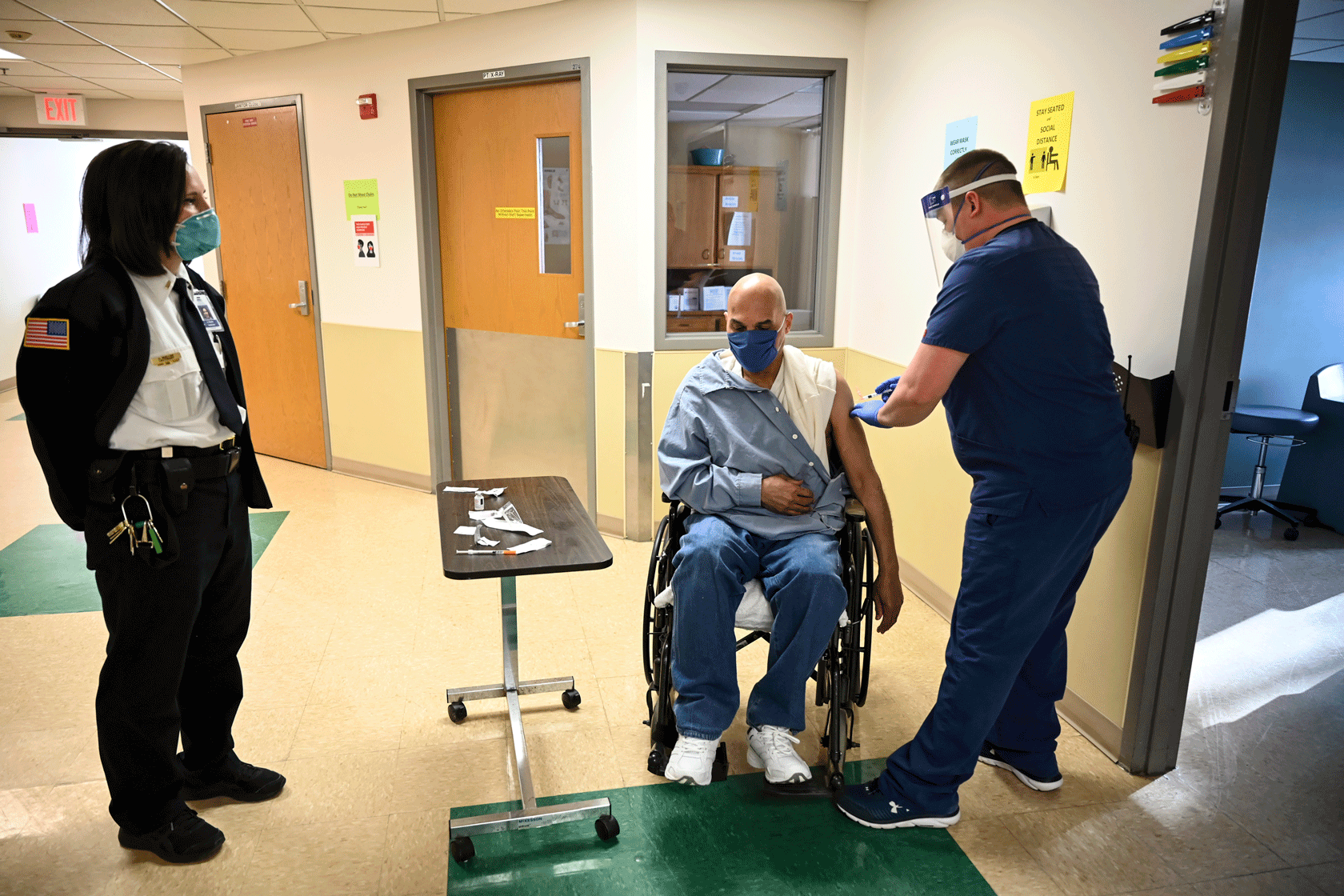 A man in a wheelchair is seen receiving a vaccination in a hallway while a security guard watches.