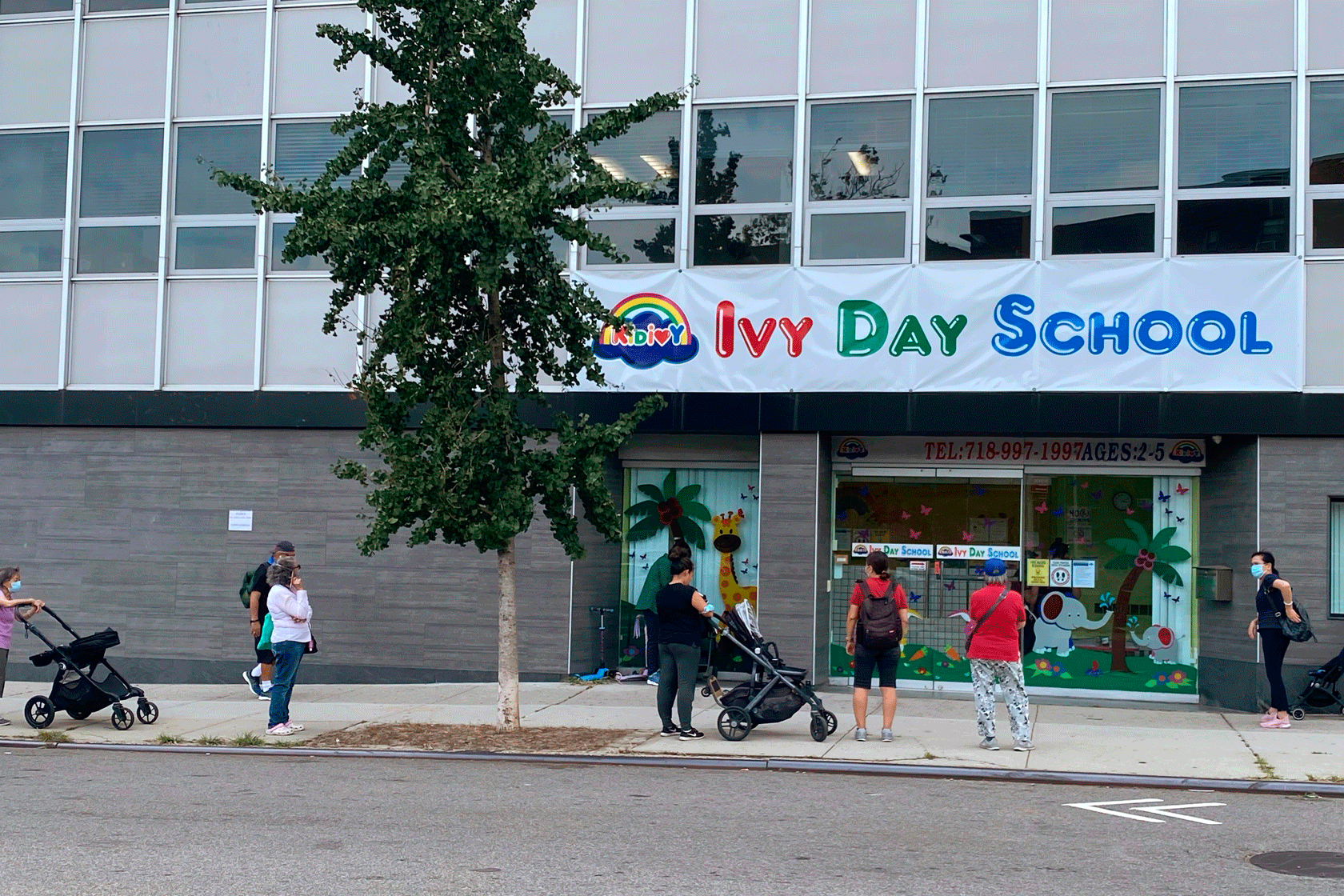 A group of parents is seen outside of a day care center. Some of them are pushing strollers.