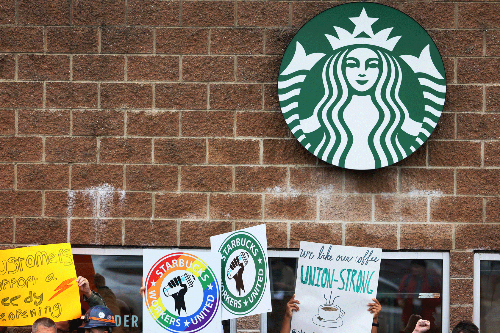 Signs in support of a union for Starbucks workers are seen in front of a brick store with the Starbucks logo on it.