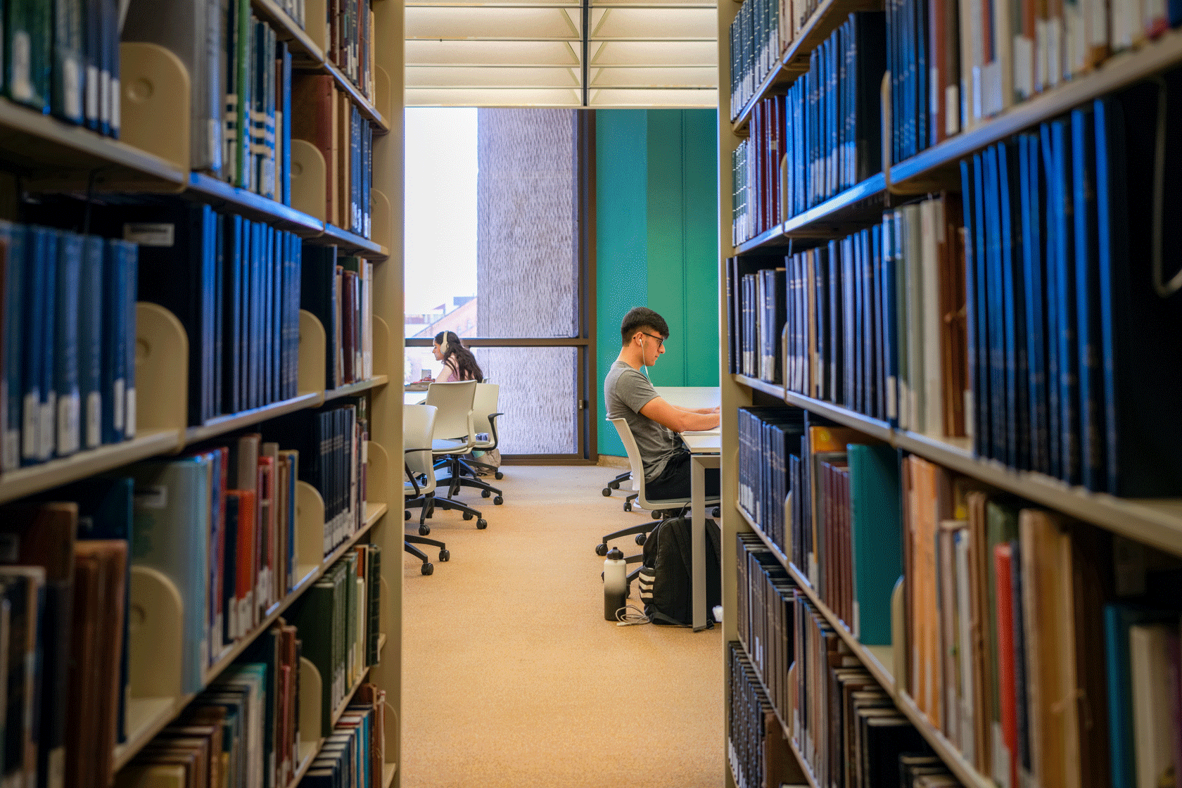 Two students are seen on their computers at tables down an aisle of books.