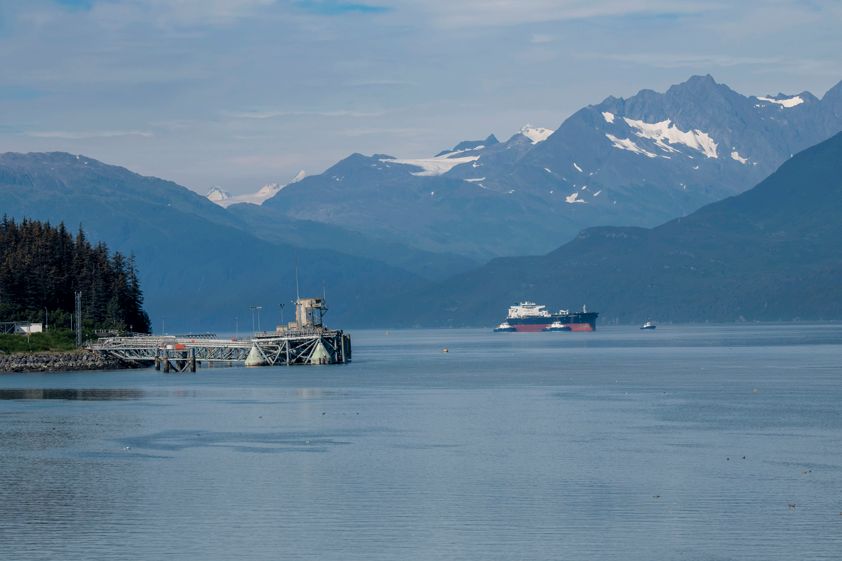A dock is seen on the left with an oil tanker on the right and mountains in the distance.