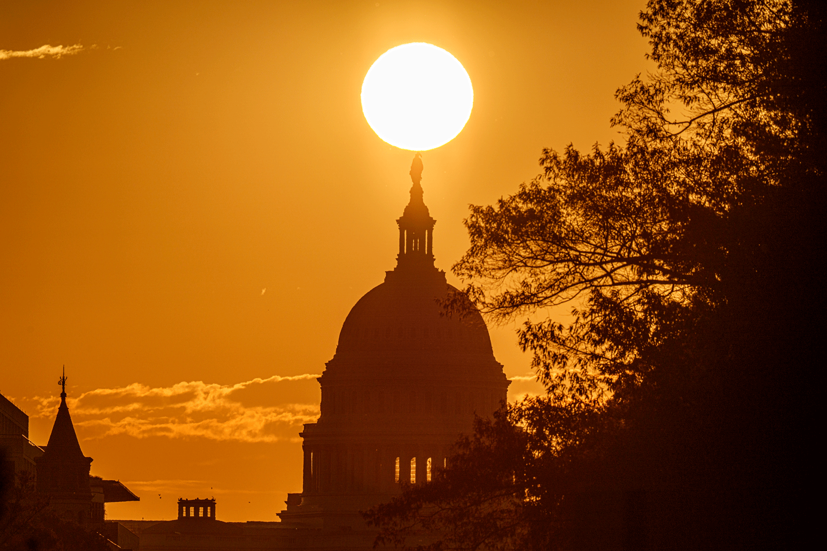 The dome of the U.S. Capitol building is seen below the sun and in front of a bright orange sky.
