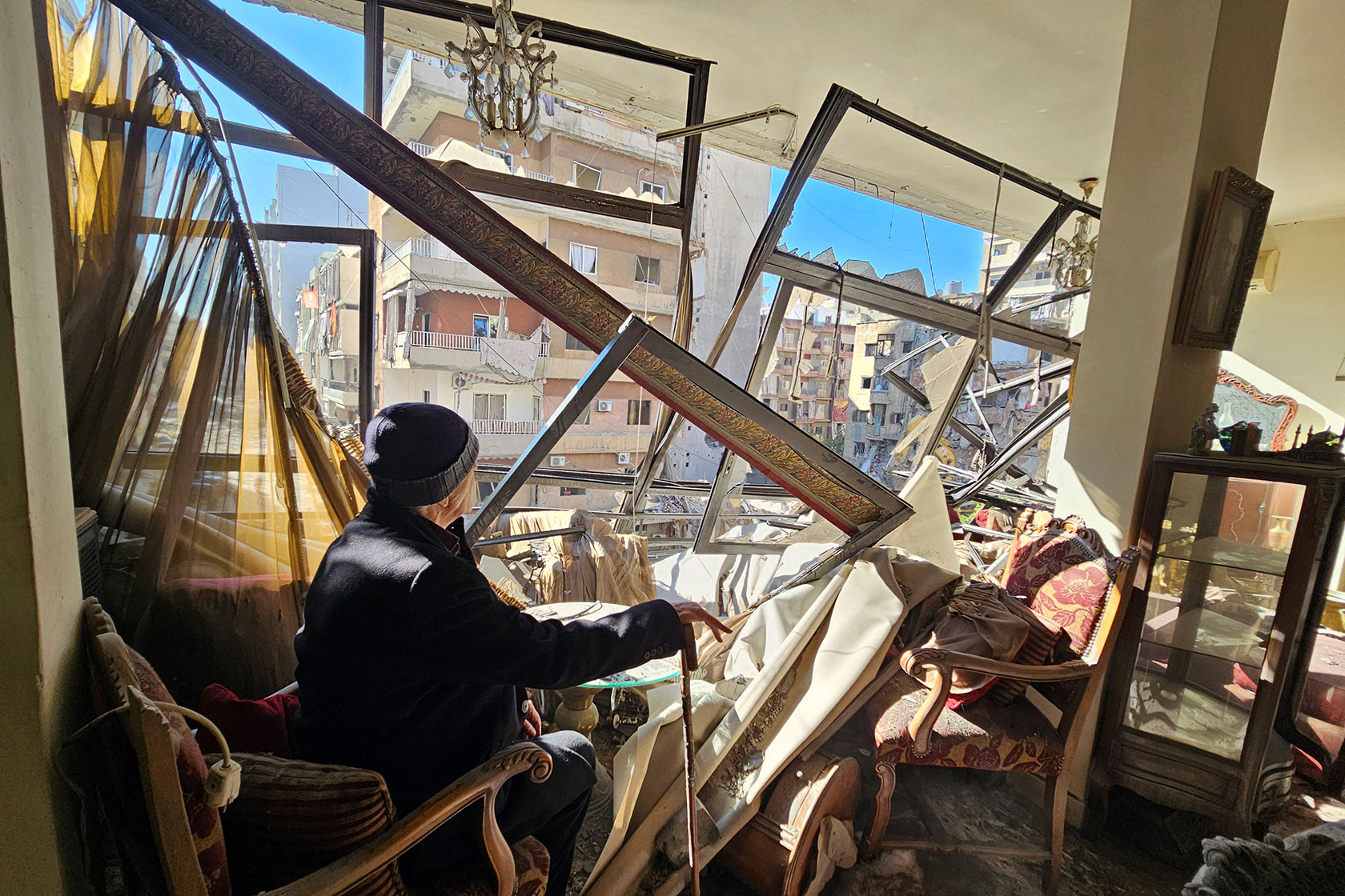 Photo shows a man sitting in a chair with his hand resting on a cane, looking out of the window of his destroyed home