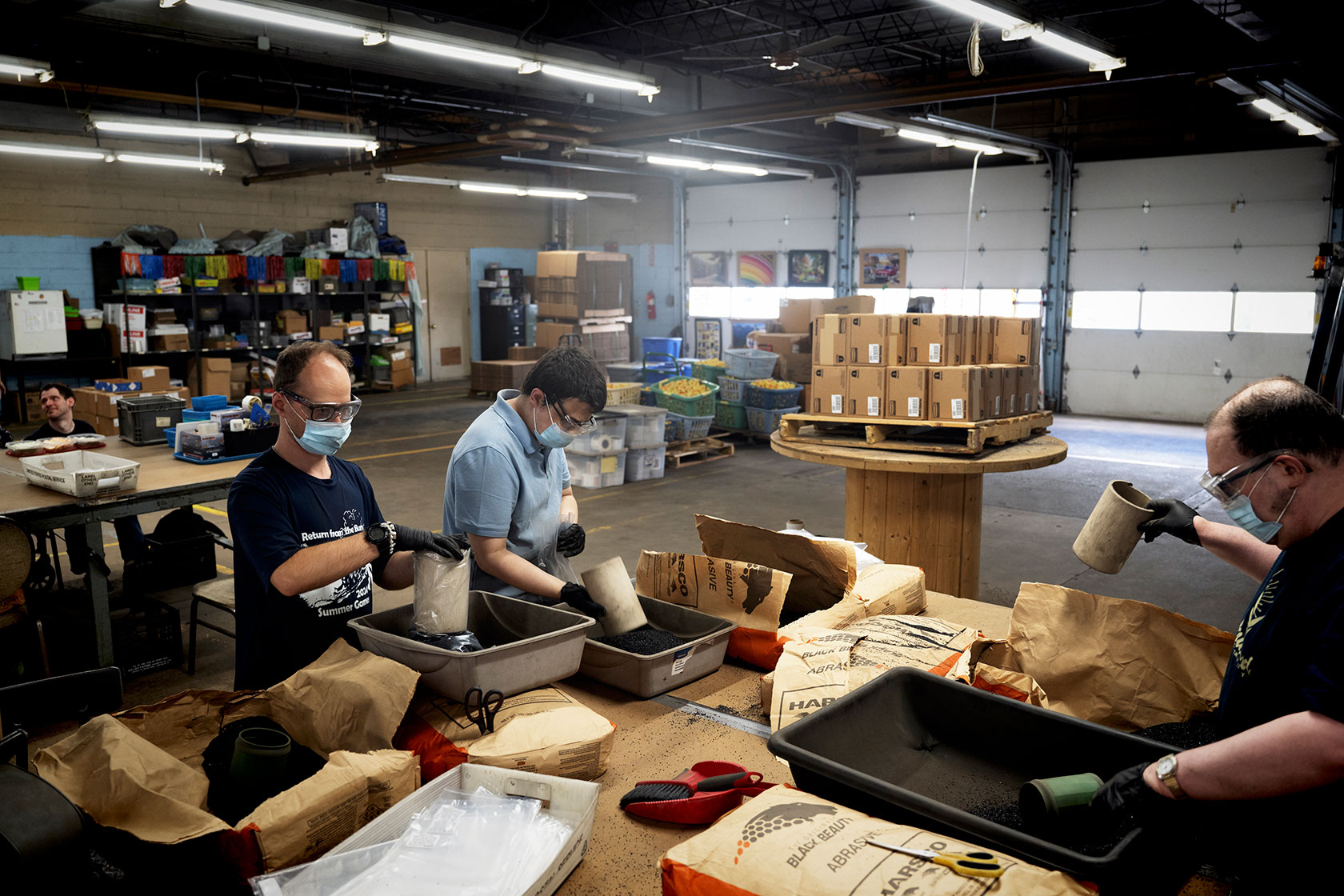 Photo shows three men wearing protective gear sorting materials in trays in a large garage warehouse