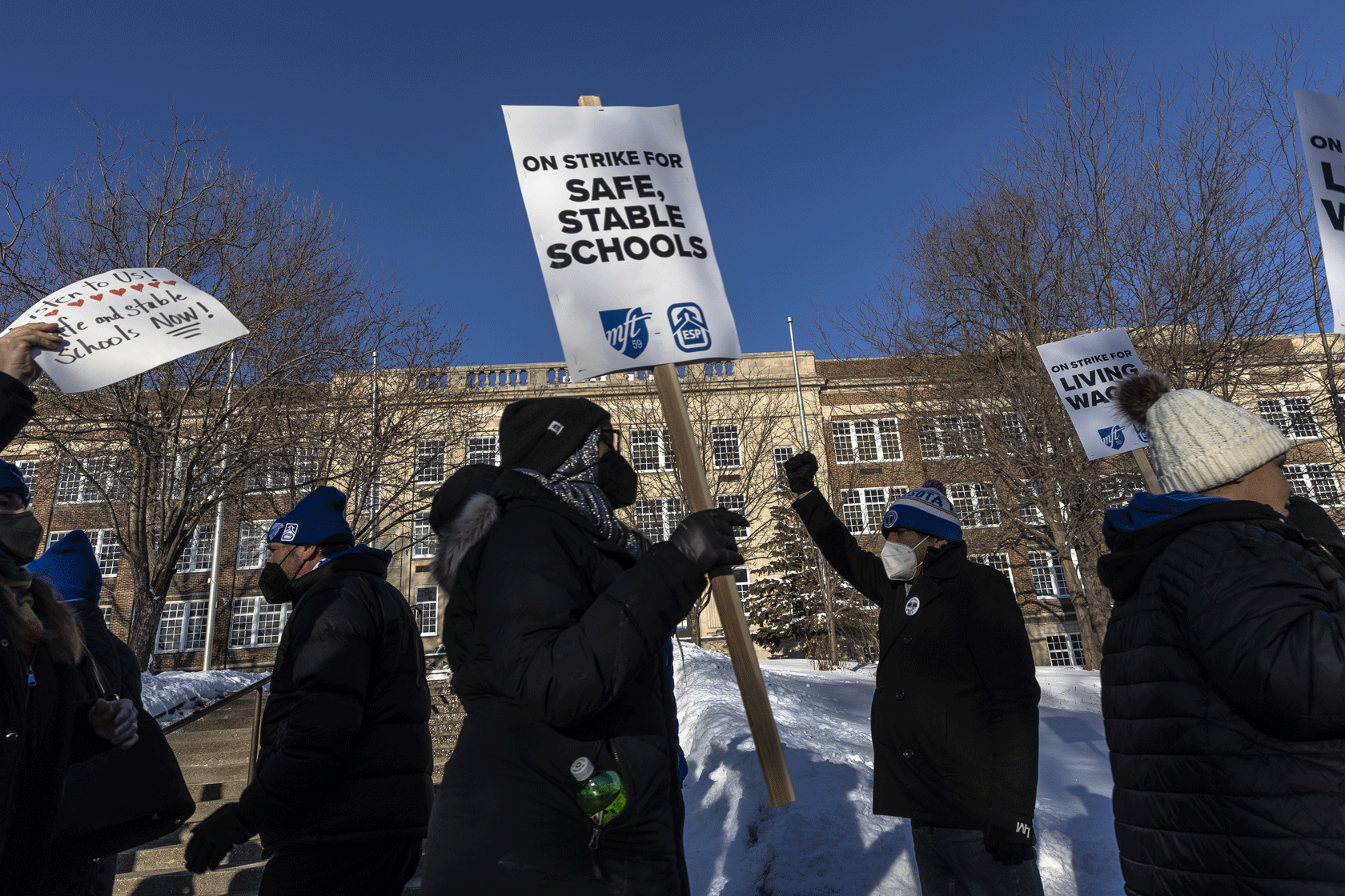 Several teachers are seen, some of whom hold placards, marching in front of a school building.