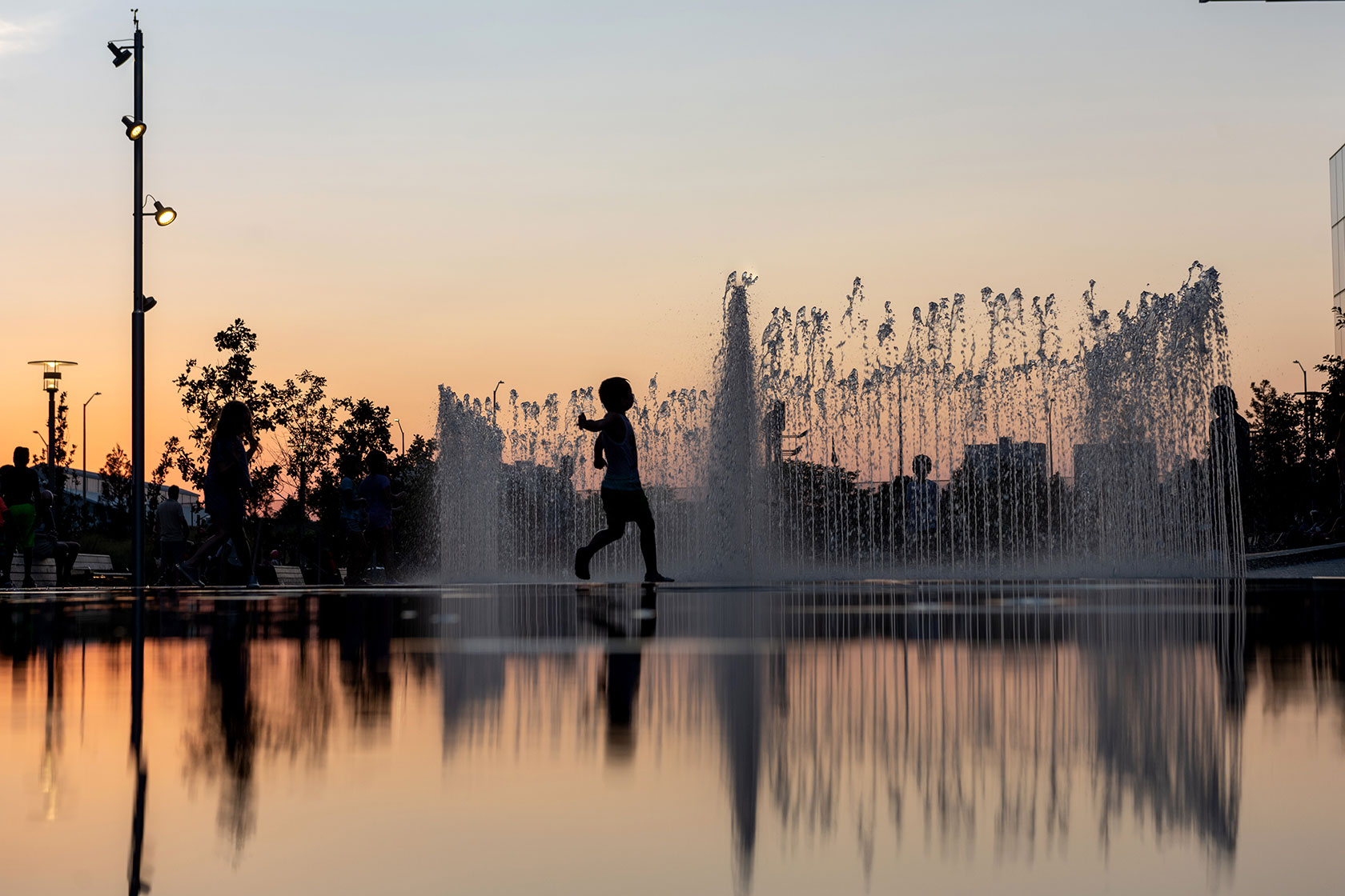 Photo shows a child silhouetted as they run through a fountain against a sunset sky