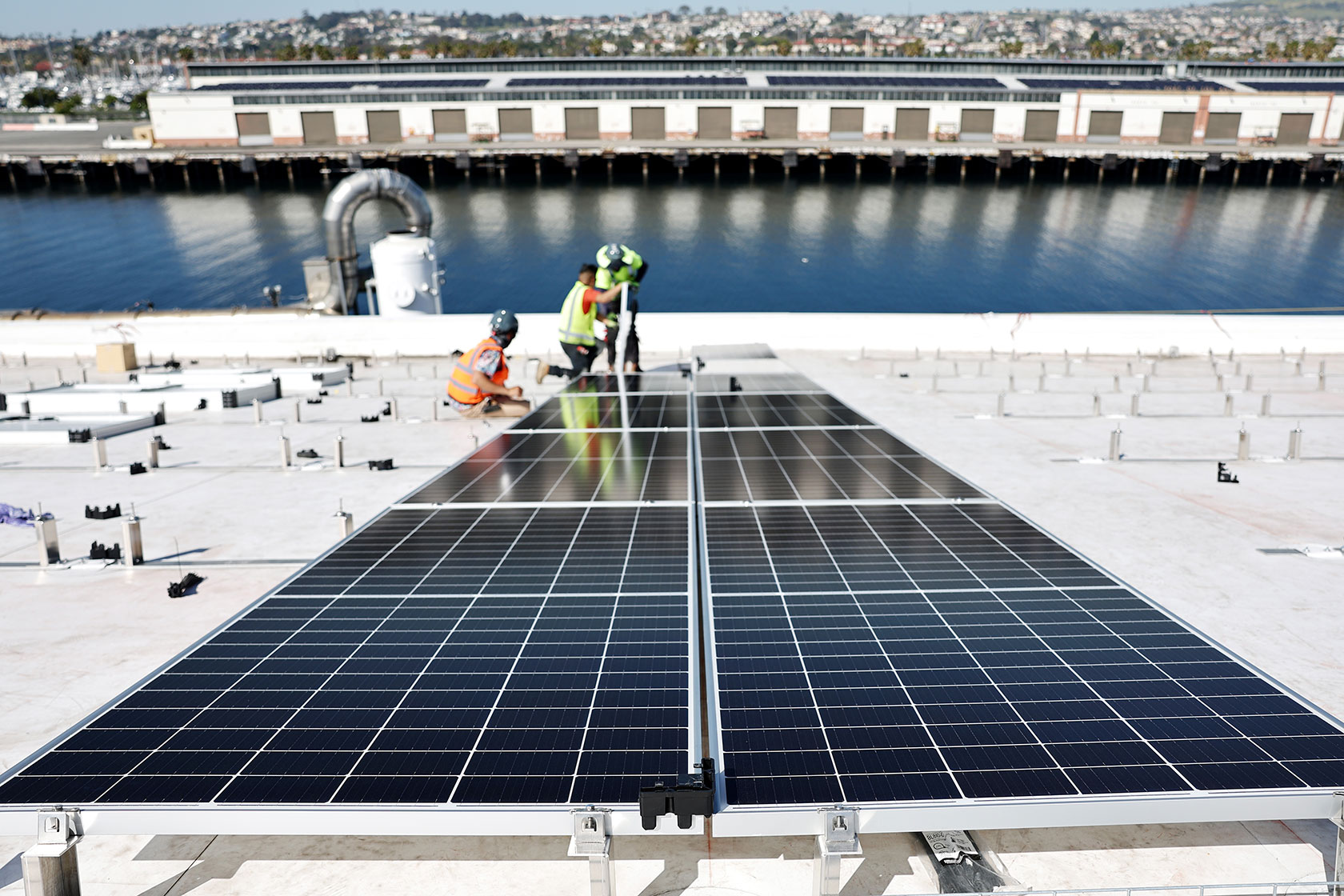 Photo shows two workers in protective gear installing a large solar panel on a while roof with a body of water in the background